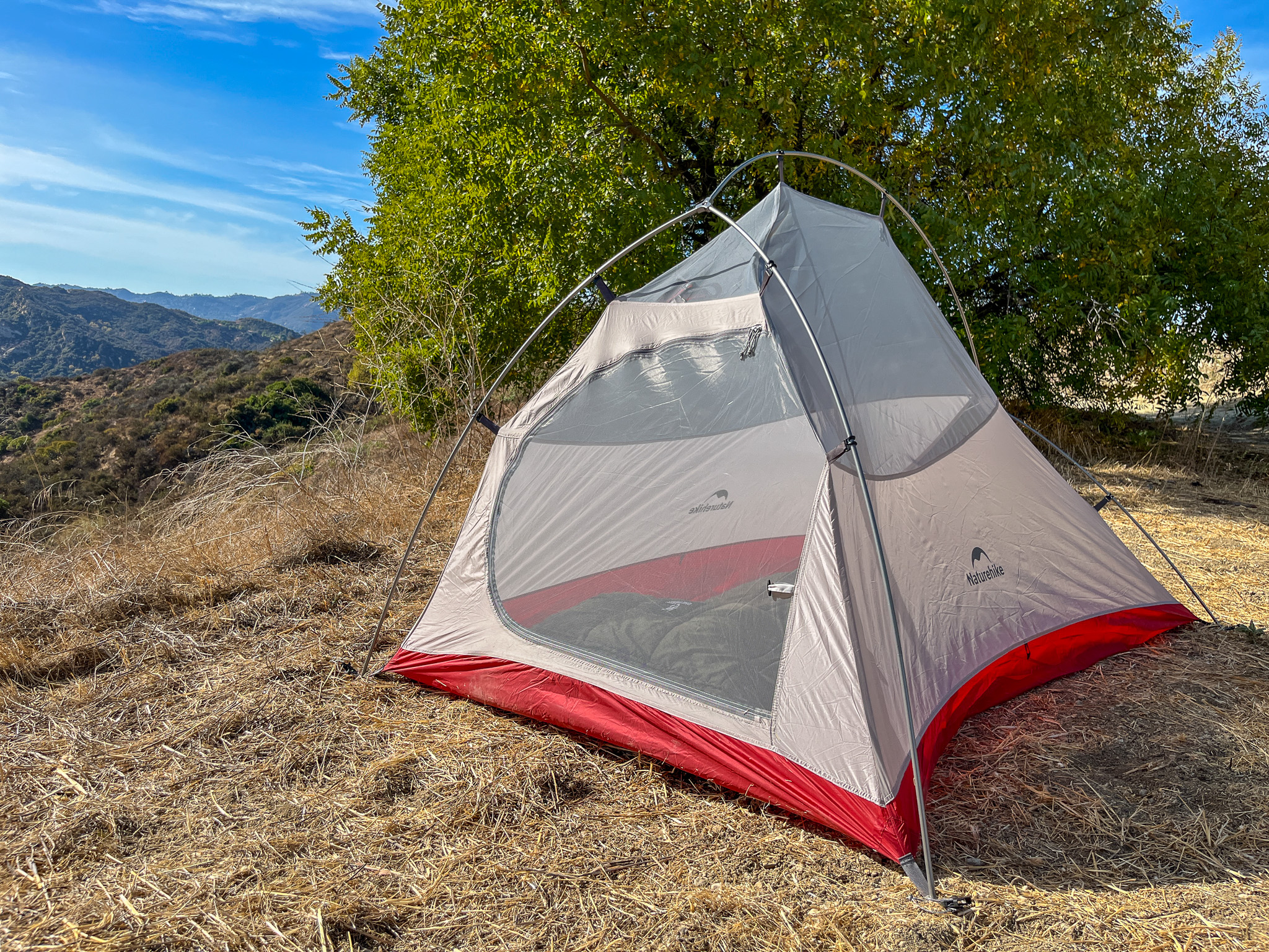 Three quarter view of the Naturehike CloudUp 2 pitched with no rainfly. It's in a scenic location with a view of rolling hills and a tree.