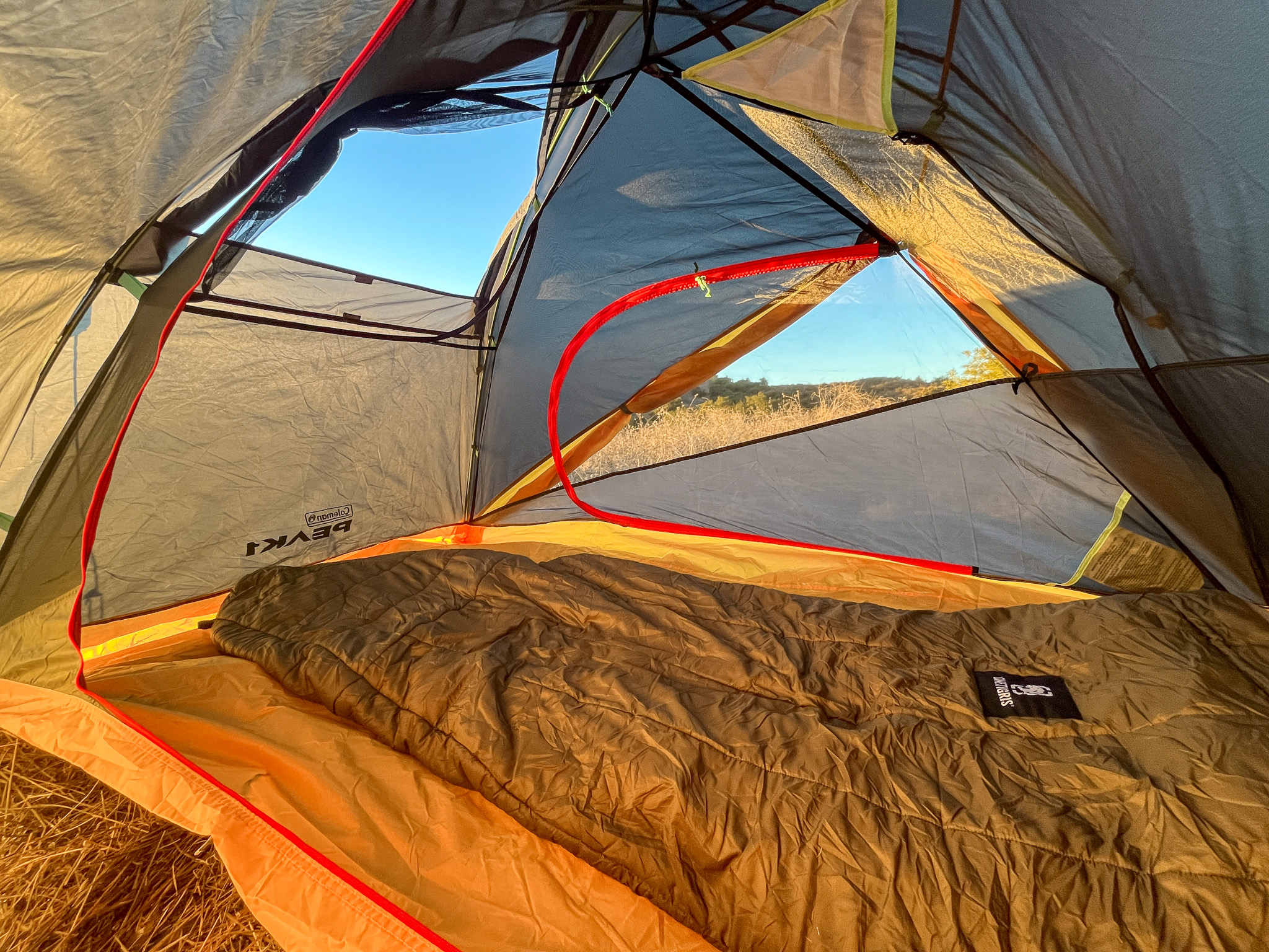 Interior view of the Coleman Peak 1 from the open doorway showing the opposite door zipped up, open stargazing panel, pockets and gear loft, and a sleeping bag. Hills and grass viewable through the back window.