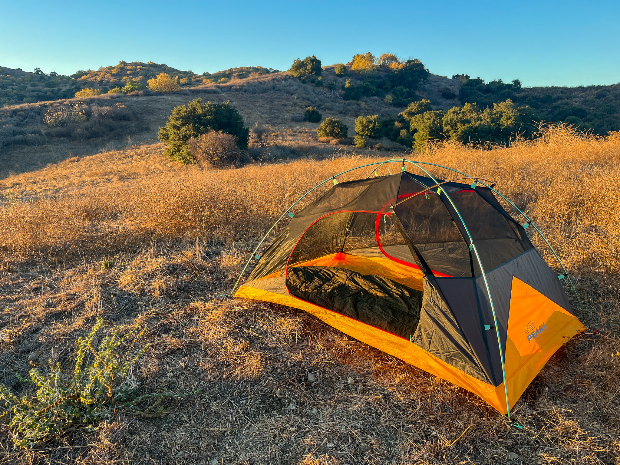 Inner mesh tent of the Coleman Peak 1 with rainfly off and one of the doors rolled back. Shows the interior space with a sleeping bag and rolling hills and bushes in the background.