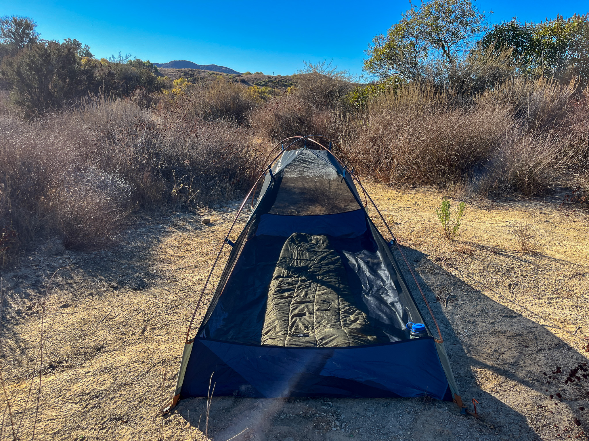 Side profile of the Kelty Late Start 2 showing the interior with a sleeping bag. surrounding hills and mountains with various grasses, bushes and trees.