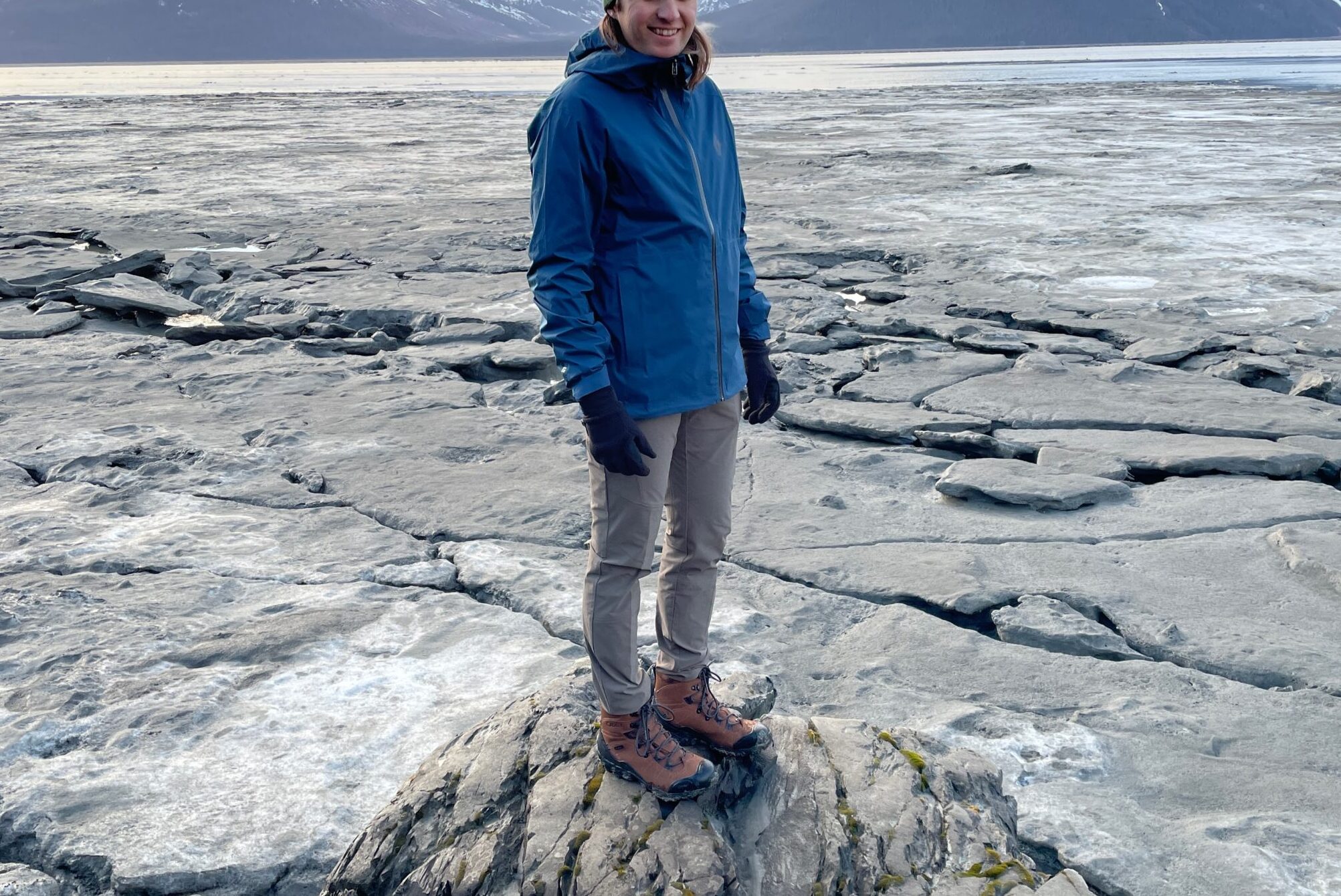 A man wearing a blue rain jacket stands along the coast.