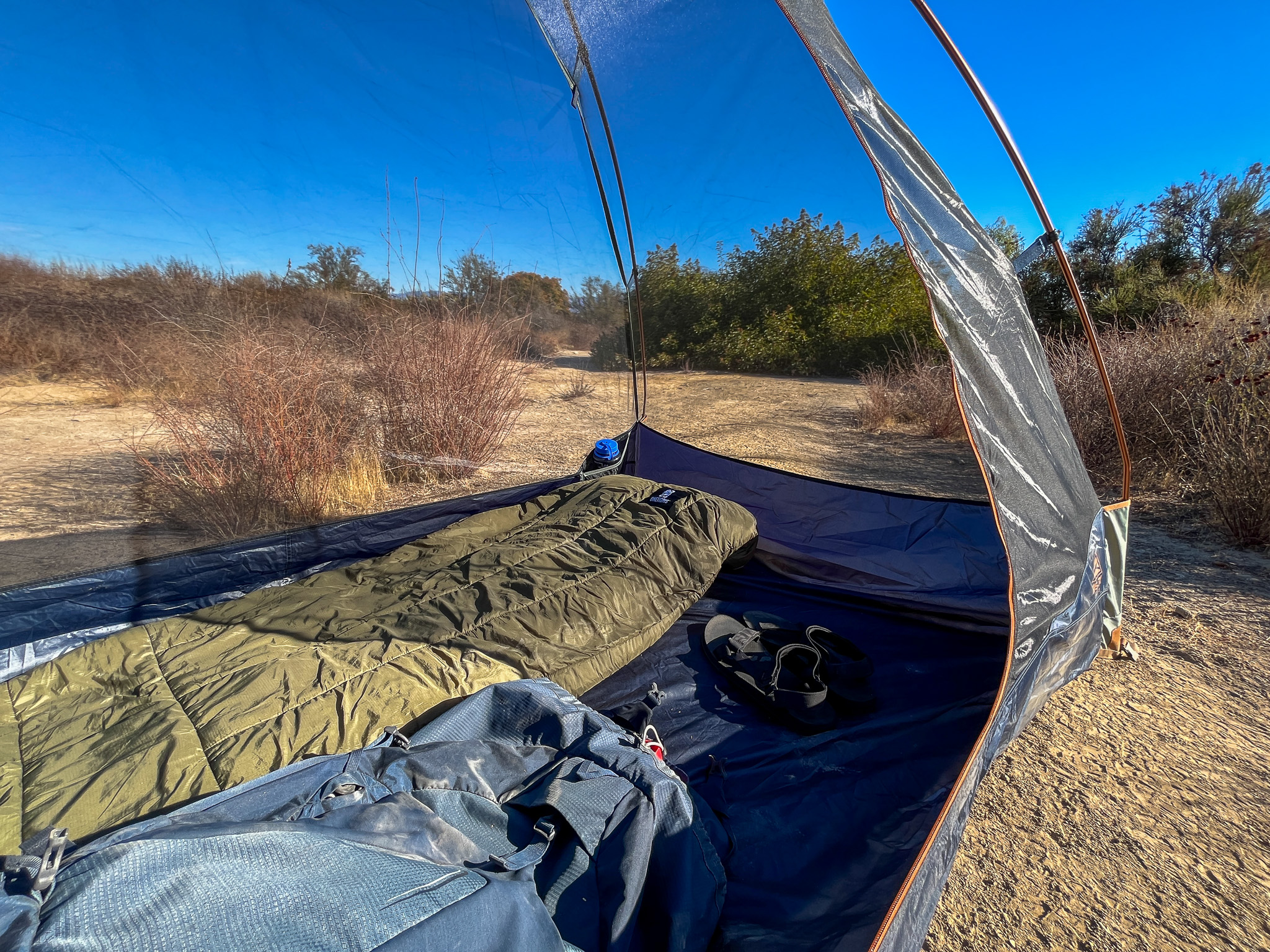 Inside view of the Kelty Late Start 2 interior showing the mesh walls, bathtub floor and floor space. Dry patches of grass and scattered bushes in the background