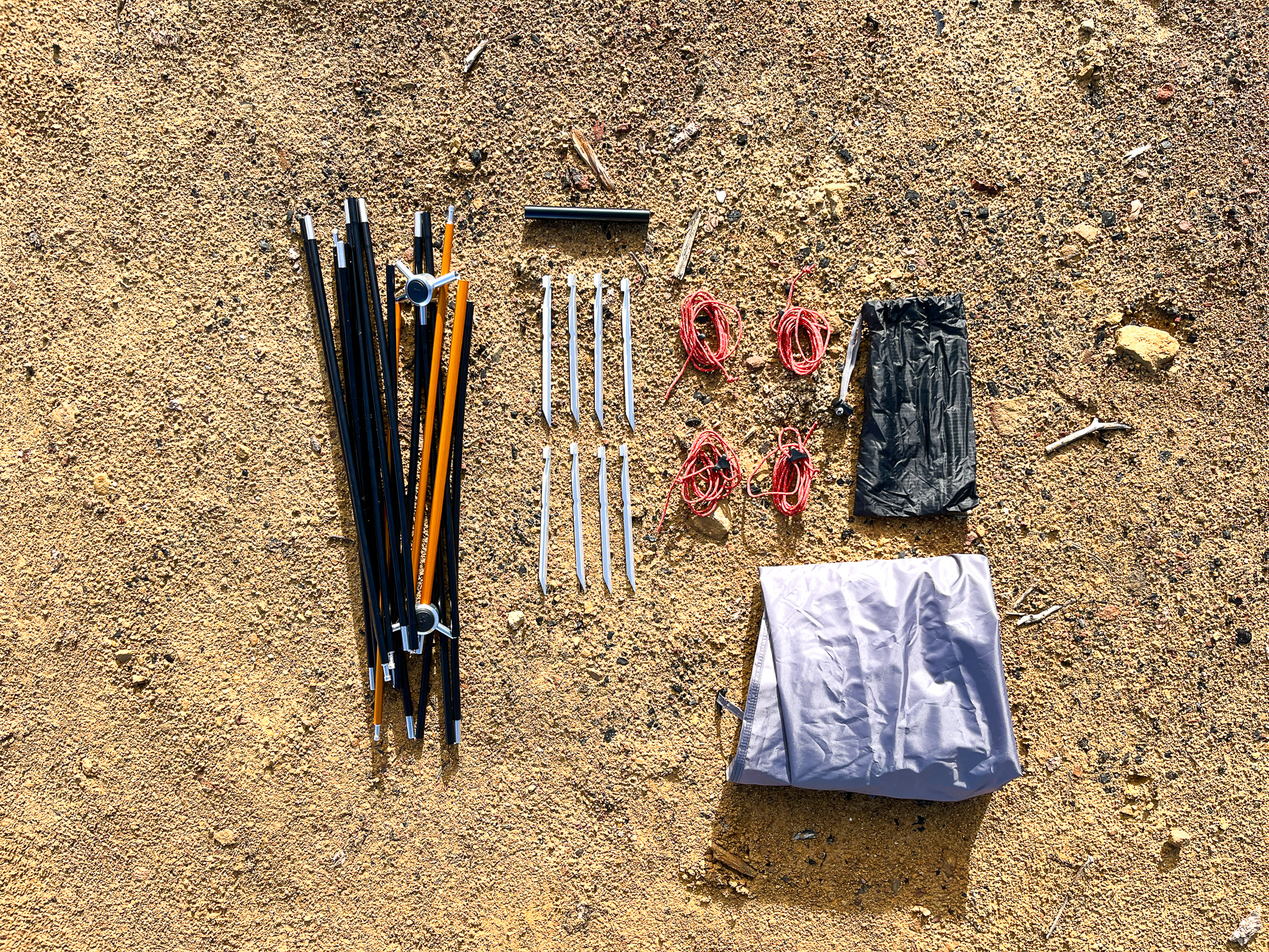 Closeup of all the REI Half Dome SL2+ hardware arranged in the dirt. This includes the hub pole system, repair splint, aluminum V-style stakes, four guy lines, a footprint, and carrying sack.