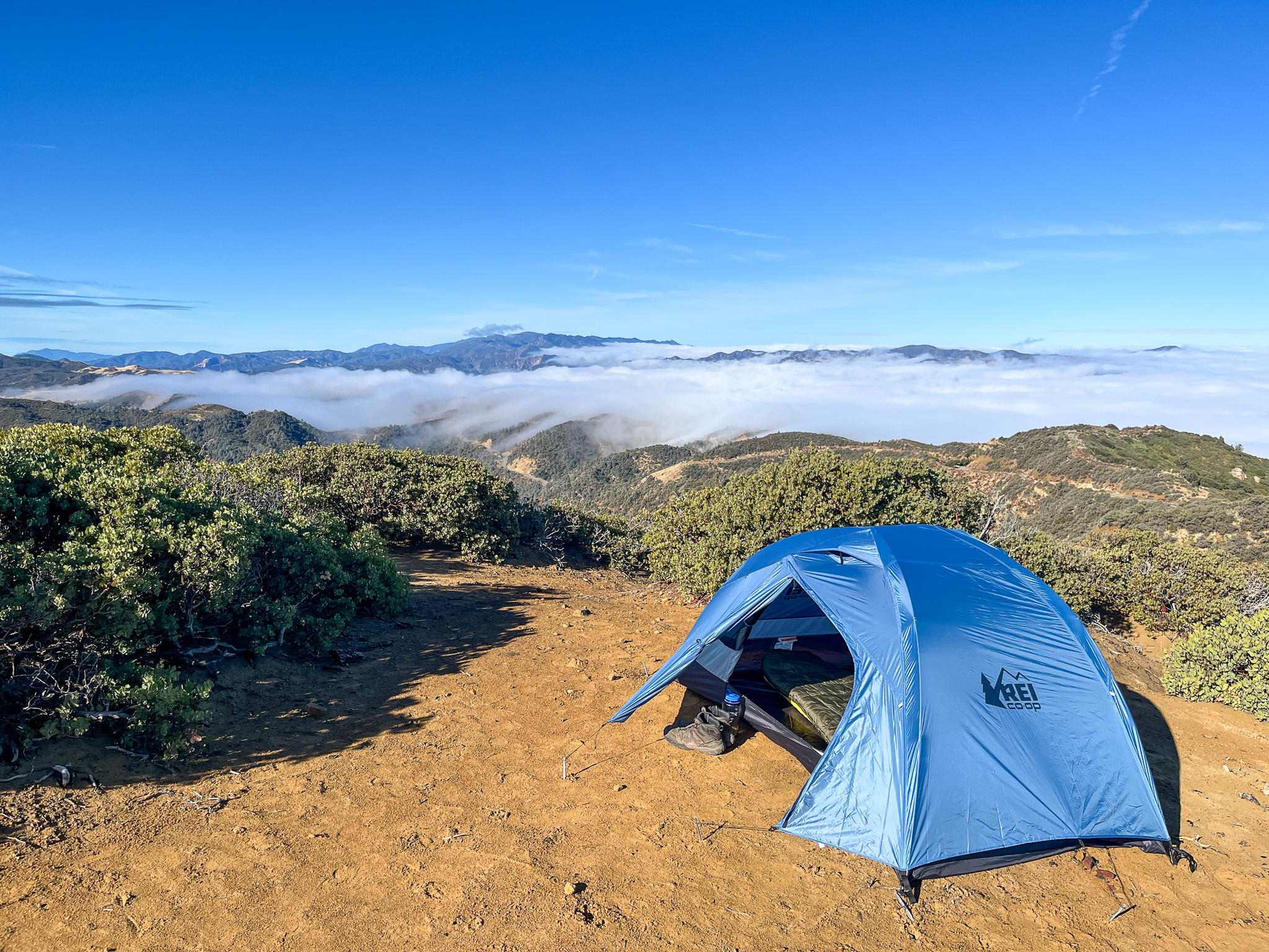 The REI Half Dome SL2+ perched on a mountainside with one door and vestibule open covering some boots and a water bottle. It overlooks a blue sky and mountain range with a cloud layer moving across.