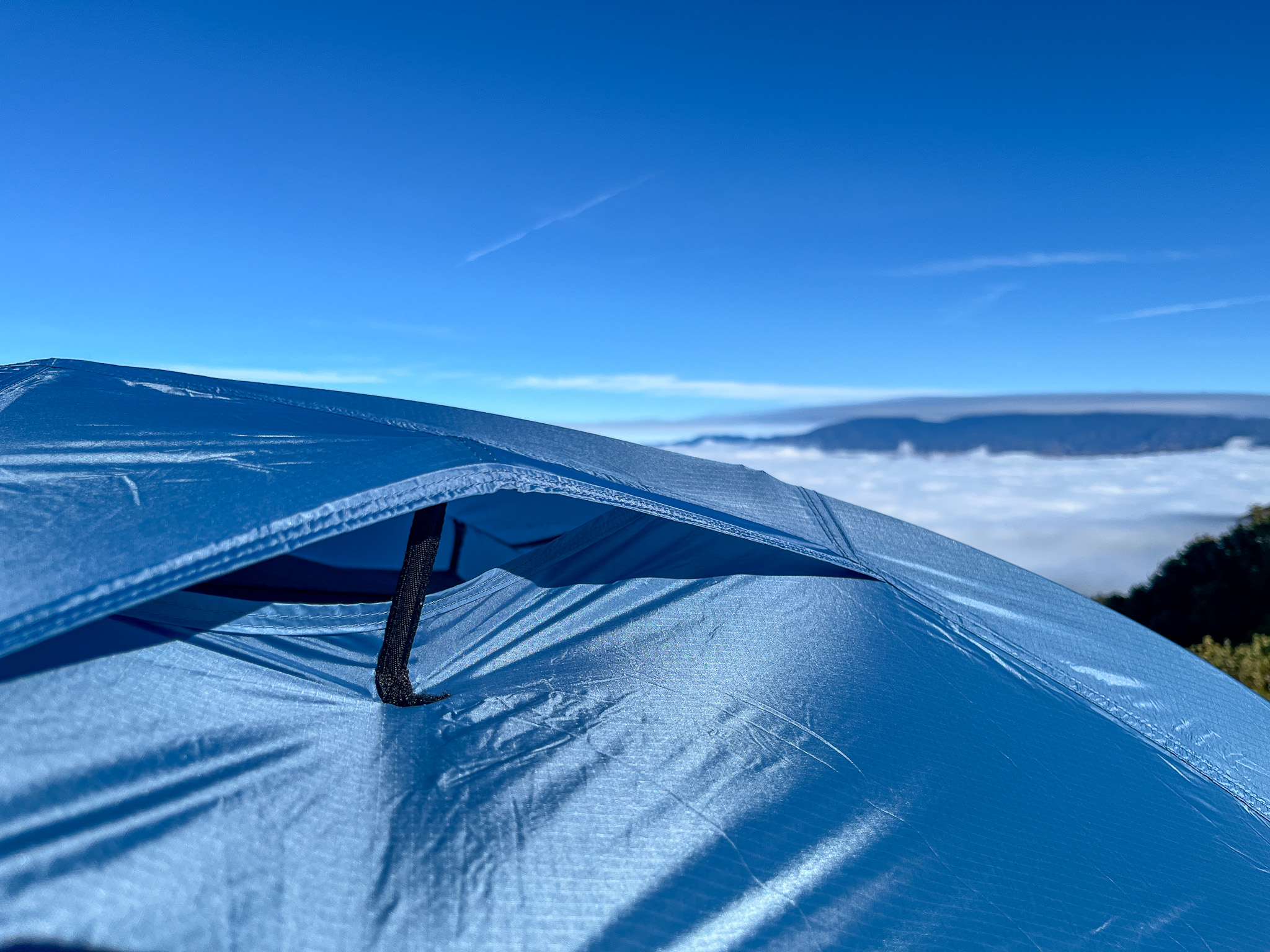 Closeup of a rainfly vent on the REI Half Dome SL2+. Shows the strutted opening, the rainfly fabric. Blue sky, mountains, and a cloud layer in the distance.