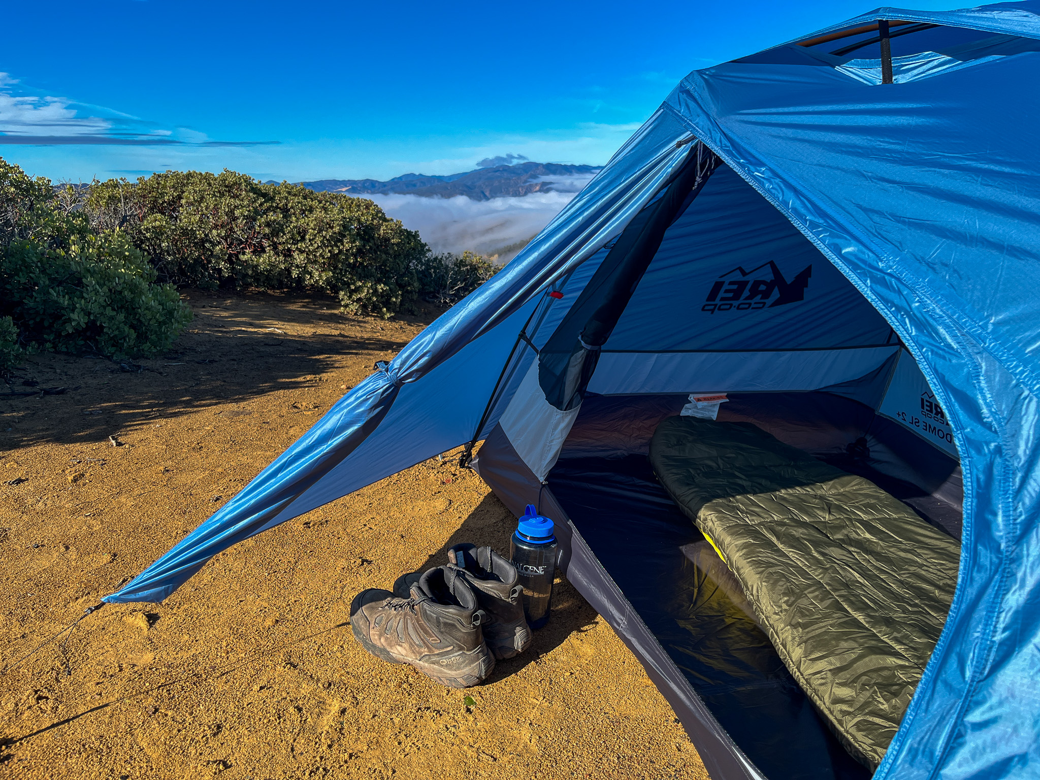 Closeup view of the REI Half Dome SL2+. Shows boots and a water bottle in the vestibule, one of the upper vents, and an open door to the interior with mountains and clouds in the background.