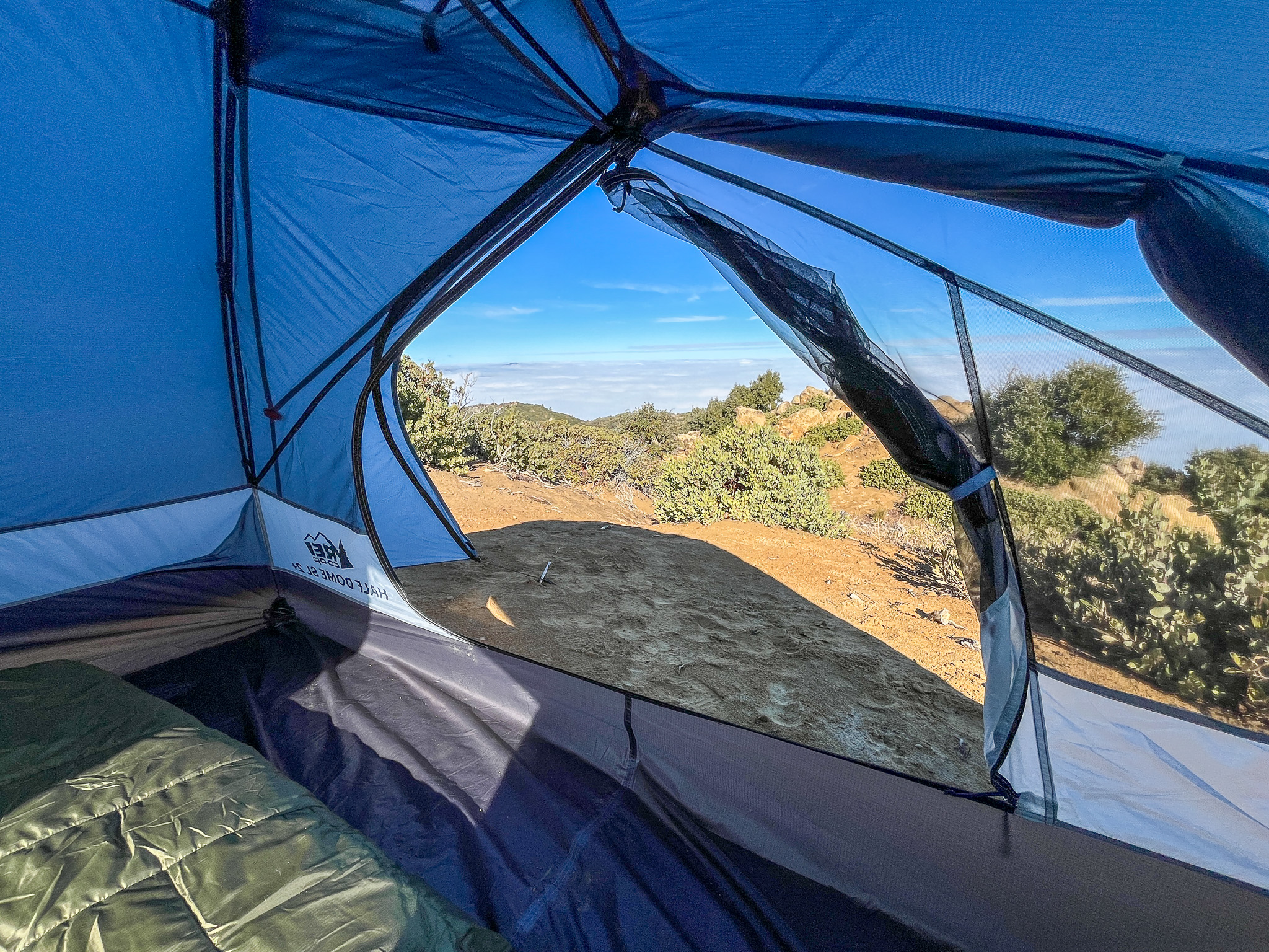 View from inside the REI Half Dome SL2+ facing out one of the doors. Shrubs, bushes, and the blue sky with a cloud layer is visible in the distance