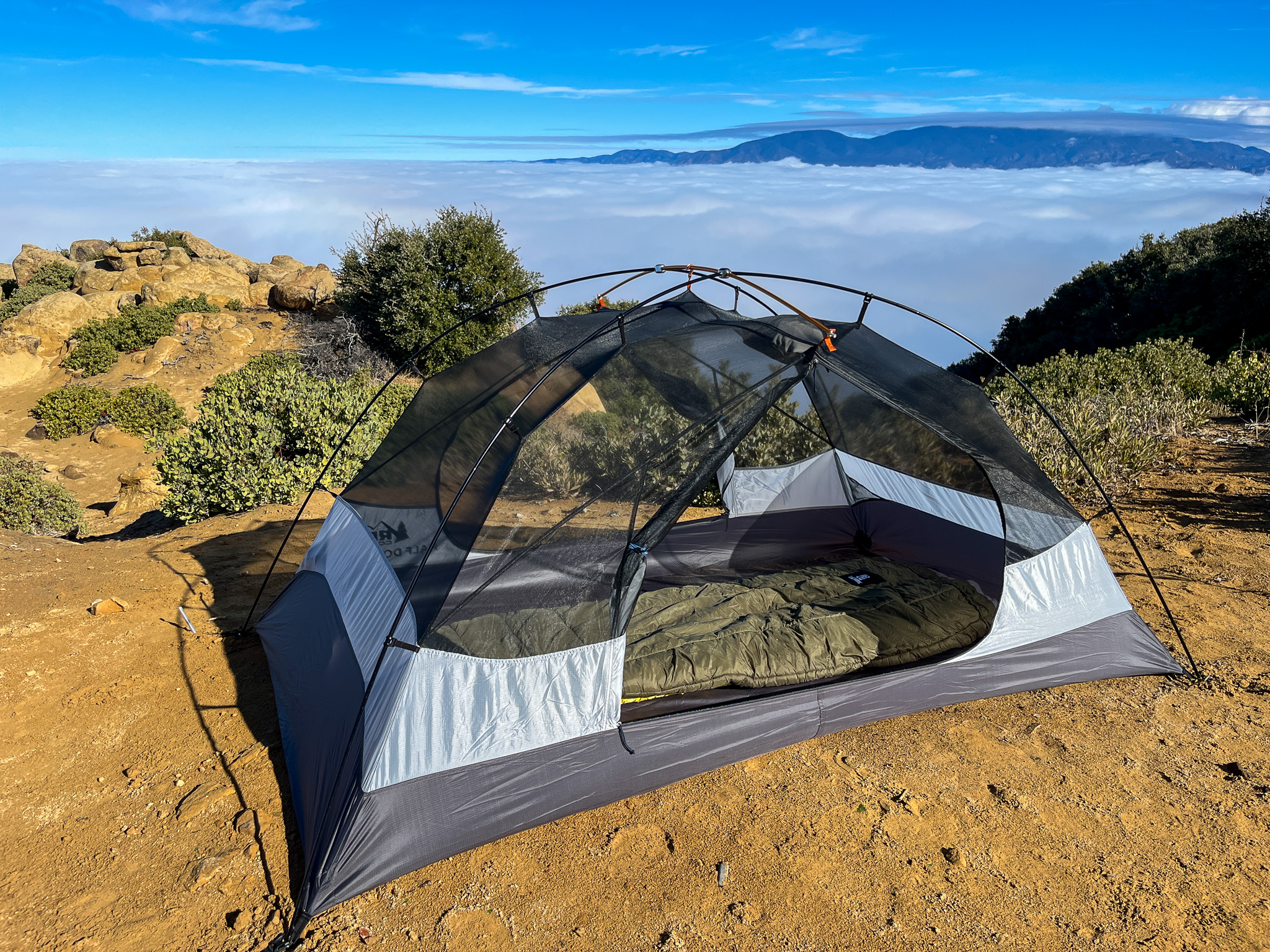 Full view of the REI Half Dome SL2+ inner tent with doors open and a sleeping bag inside. In the background is a few rocks and bushes plus large cloud layer with a mountain peeking out of it in the distance.