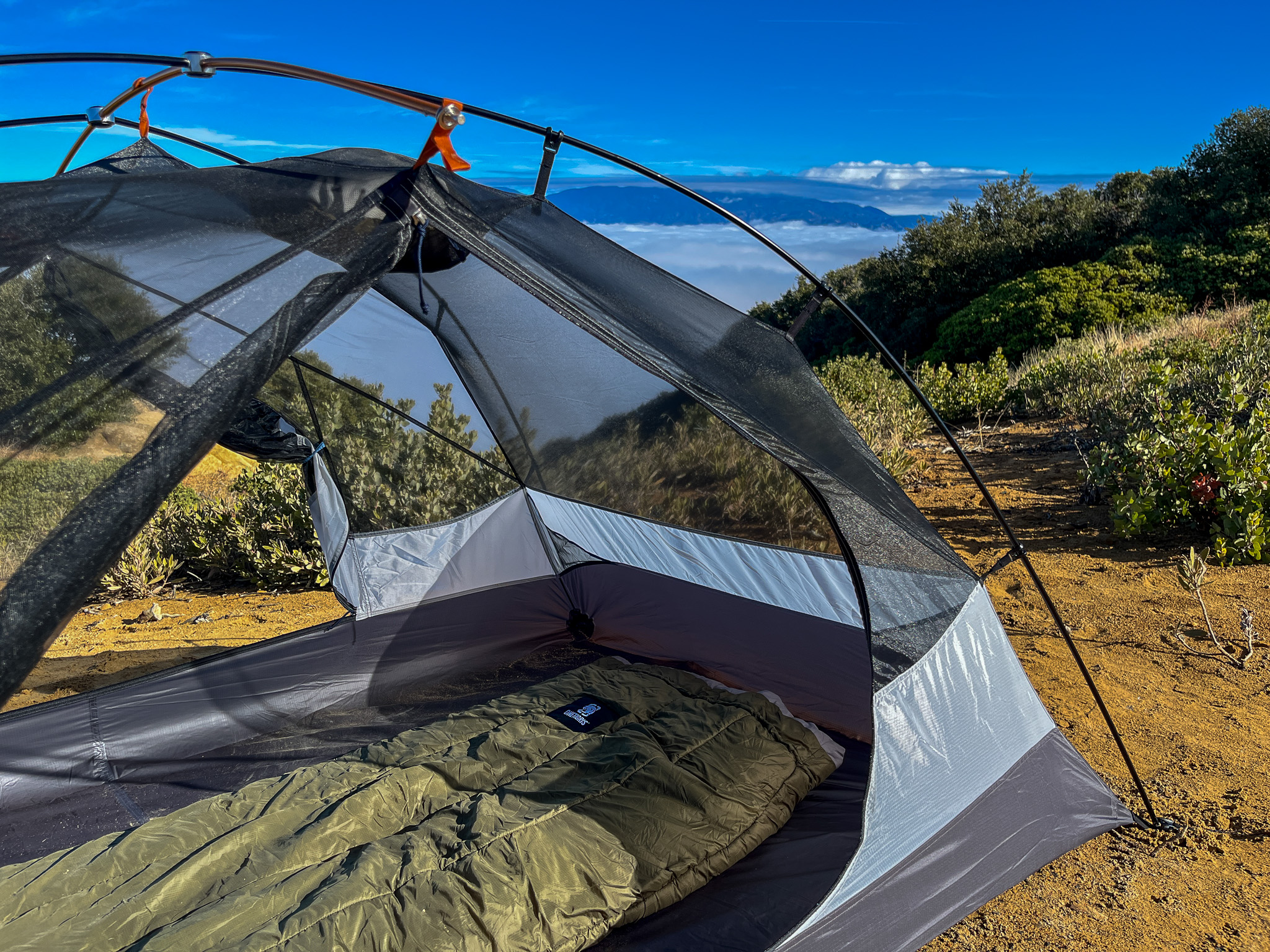 Partial interior view of the REI Half Dome SL2+ showing the floorspace with a sleeping bag inside and clipped pole. Also shows mountains and a cloud layer in the distance.