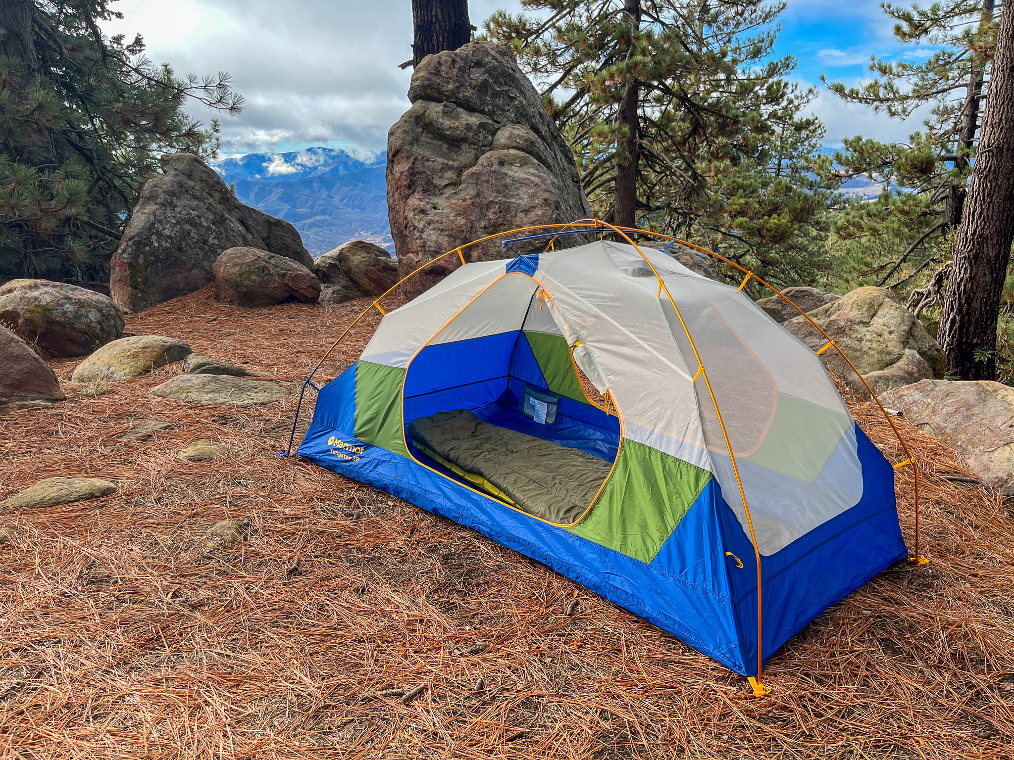 The Marmot Tungsten 2 pitched in a forest with large rocks, pine needles, and distant mountains in the background.