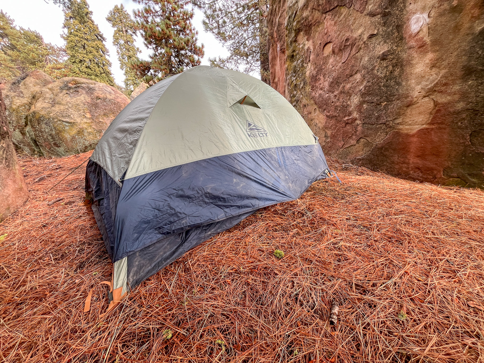 View of the backside of the Kelty Late Start 2 showing the strutted vent and pullout anchor point. Surrounded by boulders and pine trees.