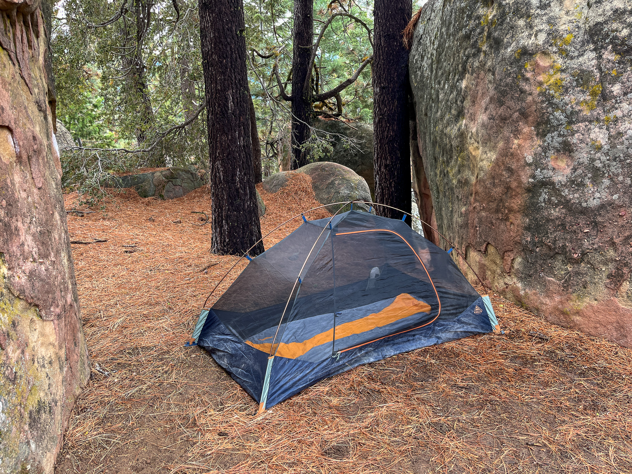 The Kelty Late Start 2 with rainfly removed and zipped up. pitched between two big boulders with surround pine forest and rocks.