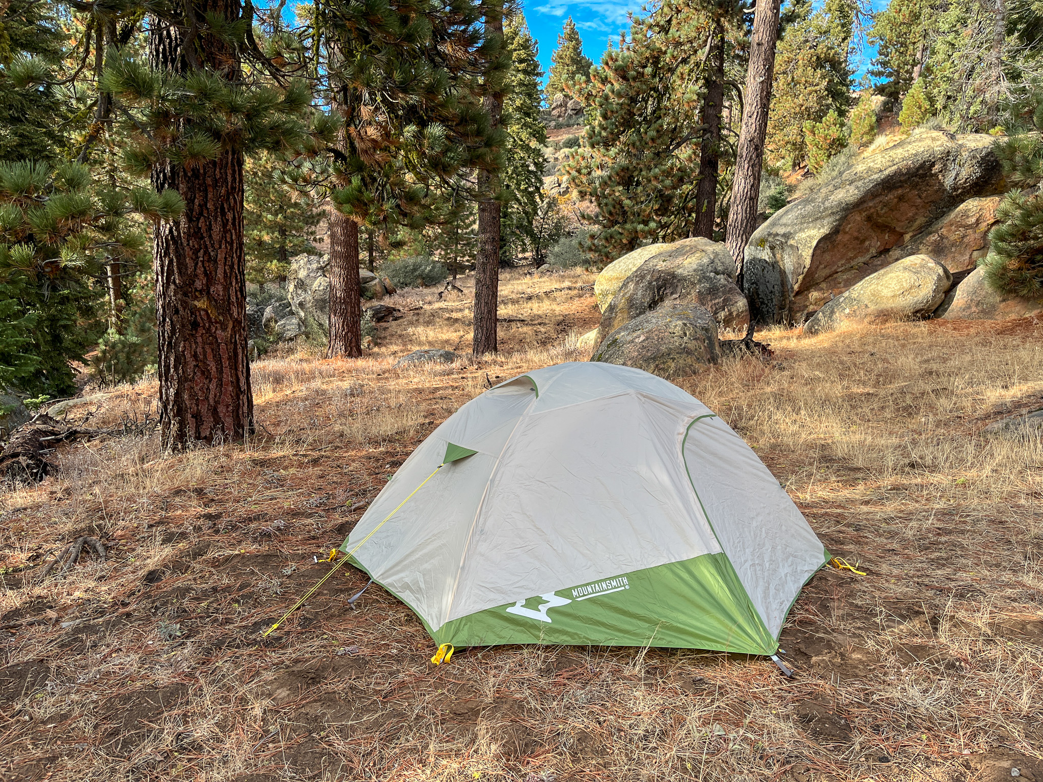 A Mountainsmith Morrison EVO 2 tent set up in a forest clearing. Tall pine trees and large granite boulders in the backdrop.