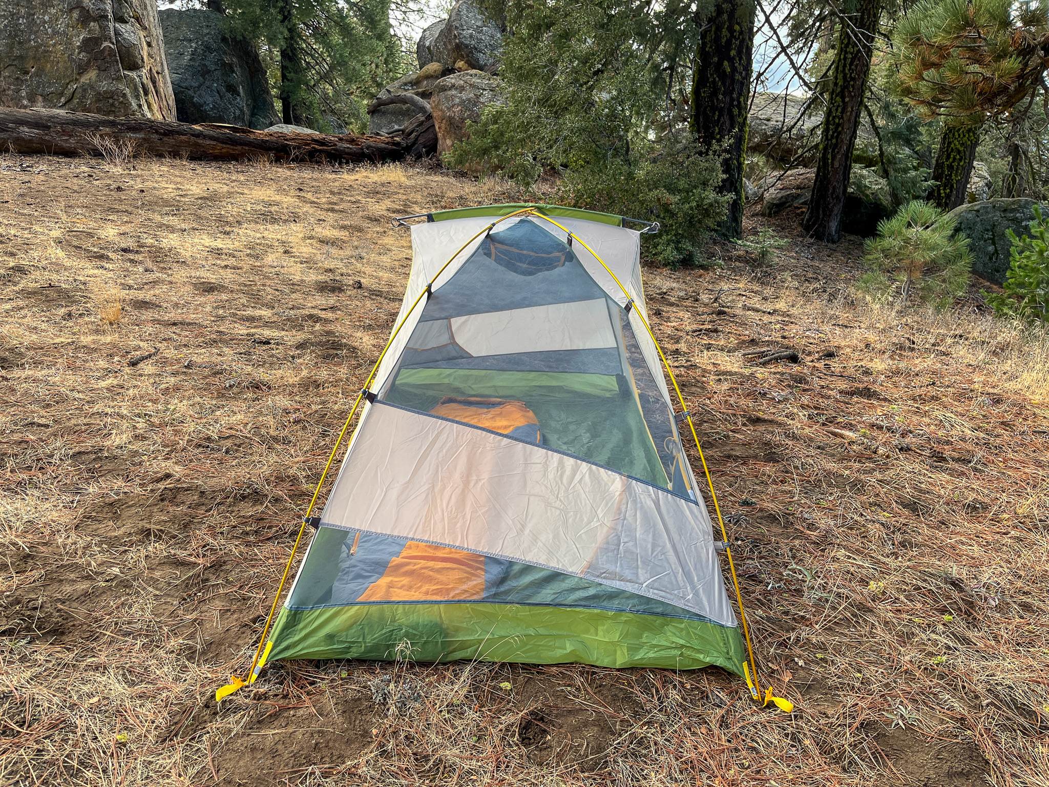 The Mountainsmith Morrison EVO 2 tent pitched in a wooded area with large boulders, trees, and a fallen log in the background.