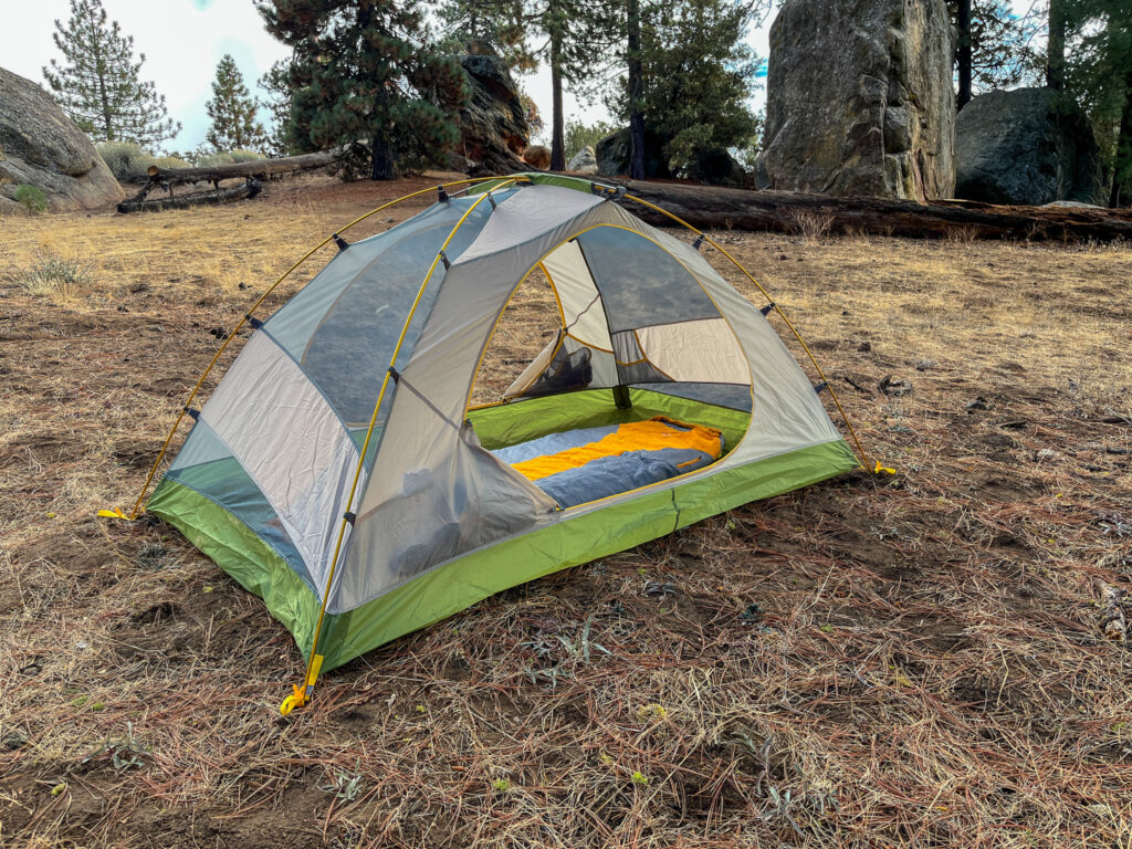 A Mountainsmith Morrison EVO 2 tent set up in the wilderness, with pine trees, fallen logs, and large boulders in the background.