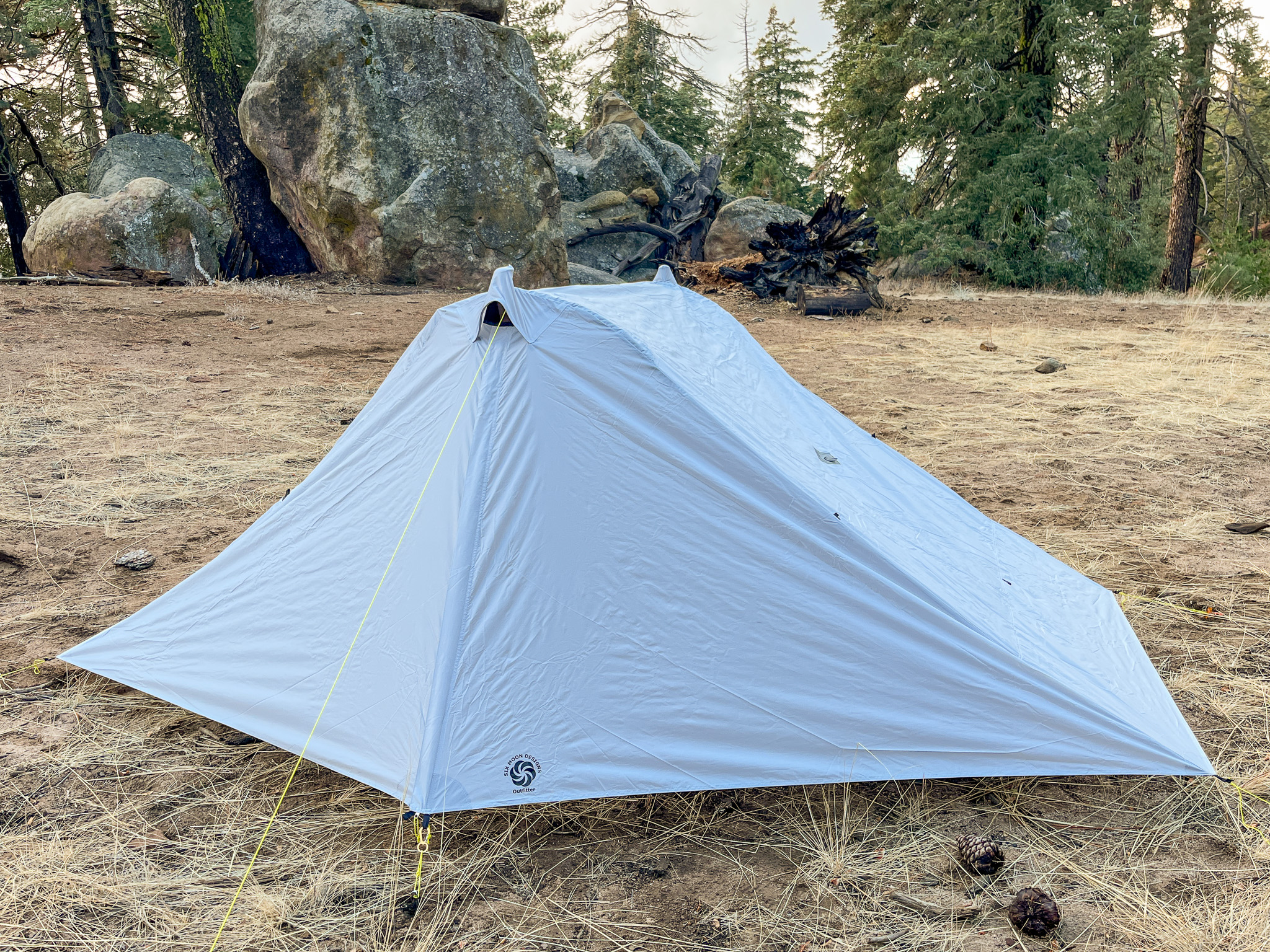 Slight three-quarter view of the Six Moon Designs Lunar Duo Outfitter with the canopy fully deployed and zipped up. It's pitched in a clearing with boulders and pine trees in the background.