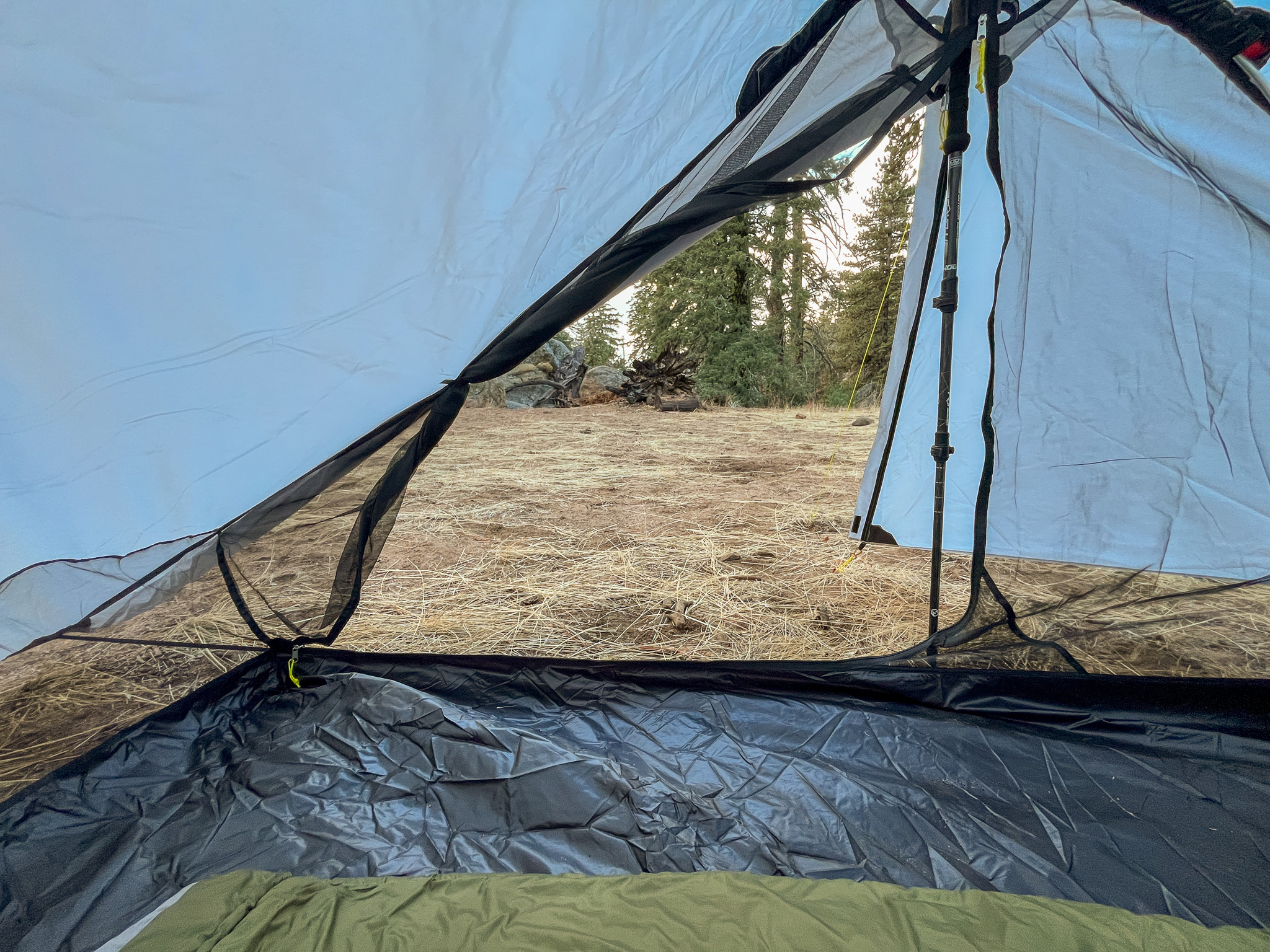 Interior view of the Six Moon Designs Lunar Duo Outfitter looking out one of the rolled back doors. A trekking pole, part of a sleeping bag, and the bathtub floor can be seen along with a pine forest visible in the background.