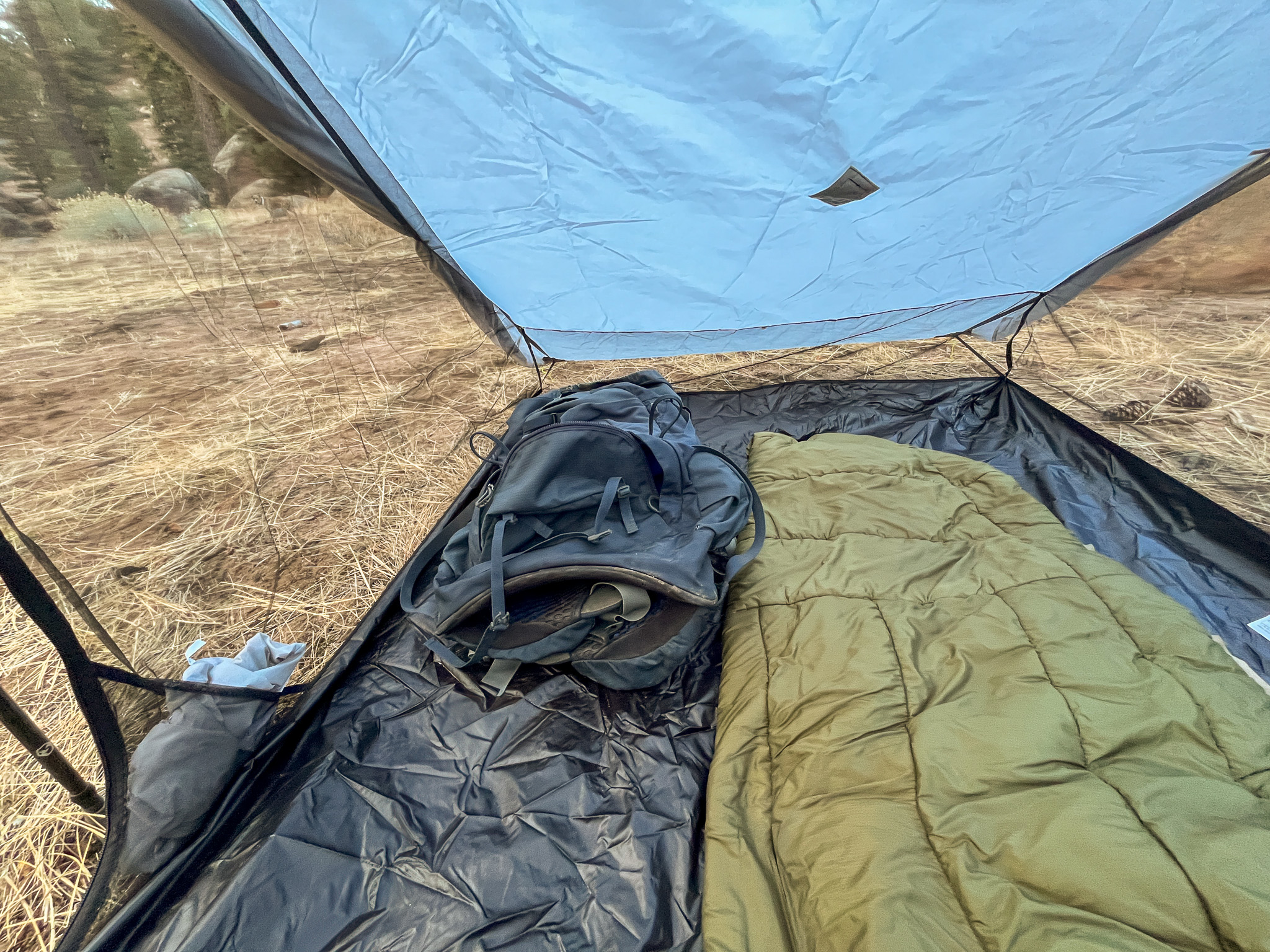 Interior view of the Six Moon Designs Lunar Duo Outfitter tent. Shows a storage pocket, backpacking pack and sleeping bag side by side. There's one canopy wall in front, and forest visible through the remaining mesh.