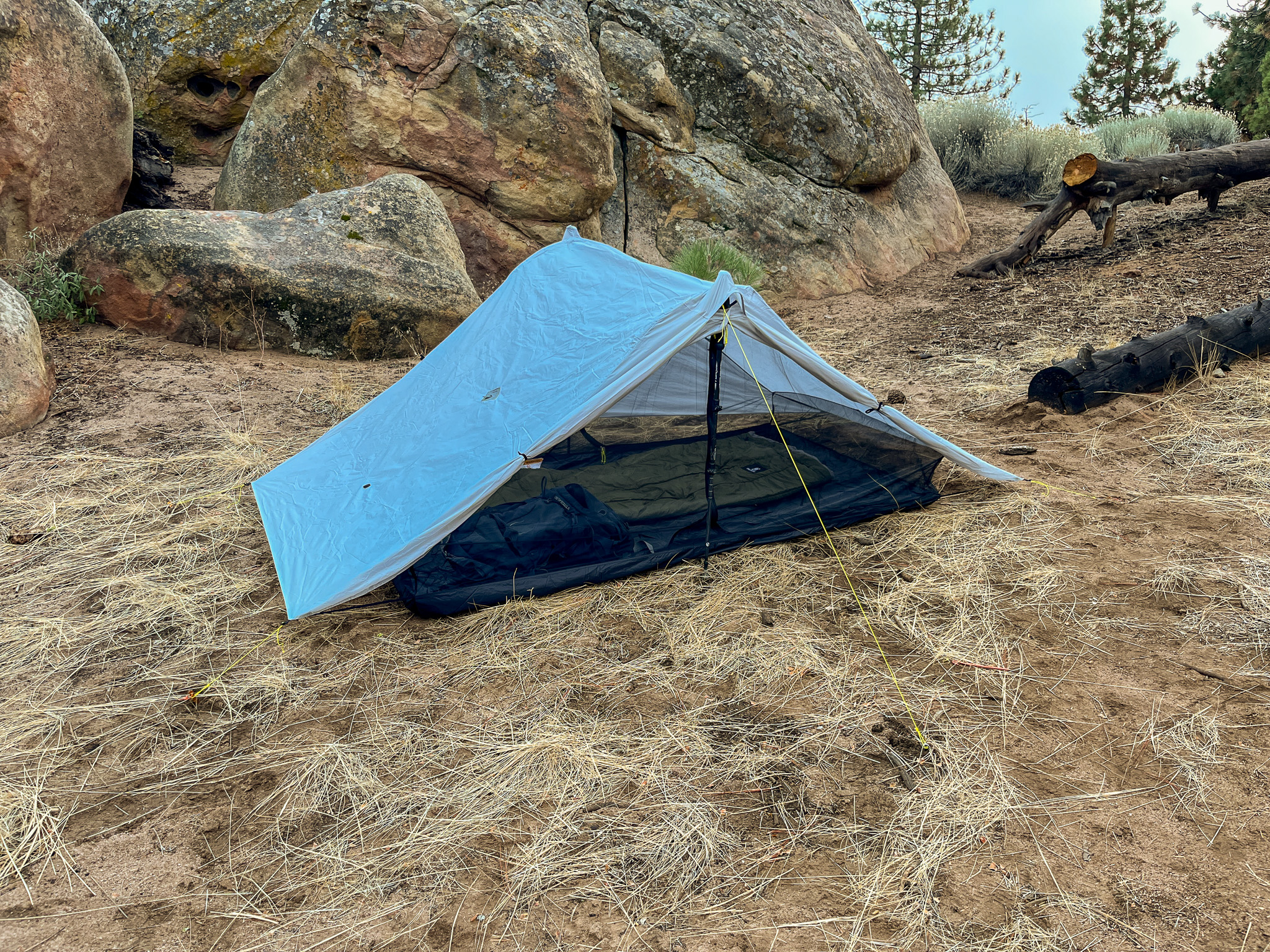 The Six Moon Designs Lunar Duo Outfitter tent pitched in front of large boulders. The vestibule doors open showing a sleeping bag and backpack inside.