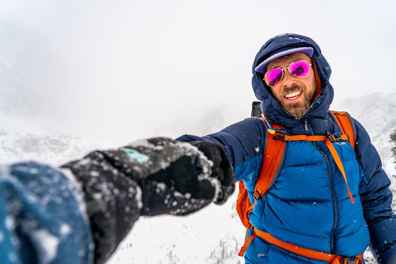 A man in winter gear and bright sunglasses fistbumps a person off camera.