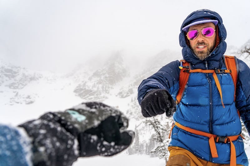 A man in winter gear and bright sunglasses fistbumps a person off camera.