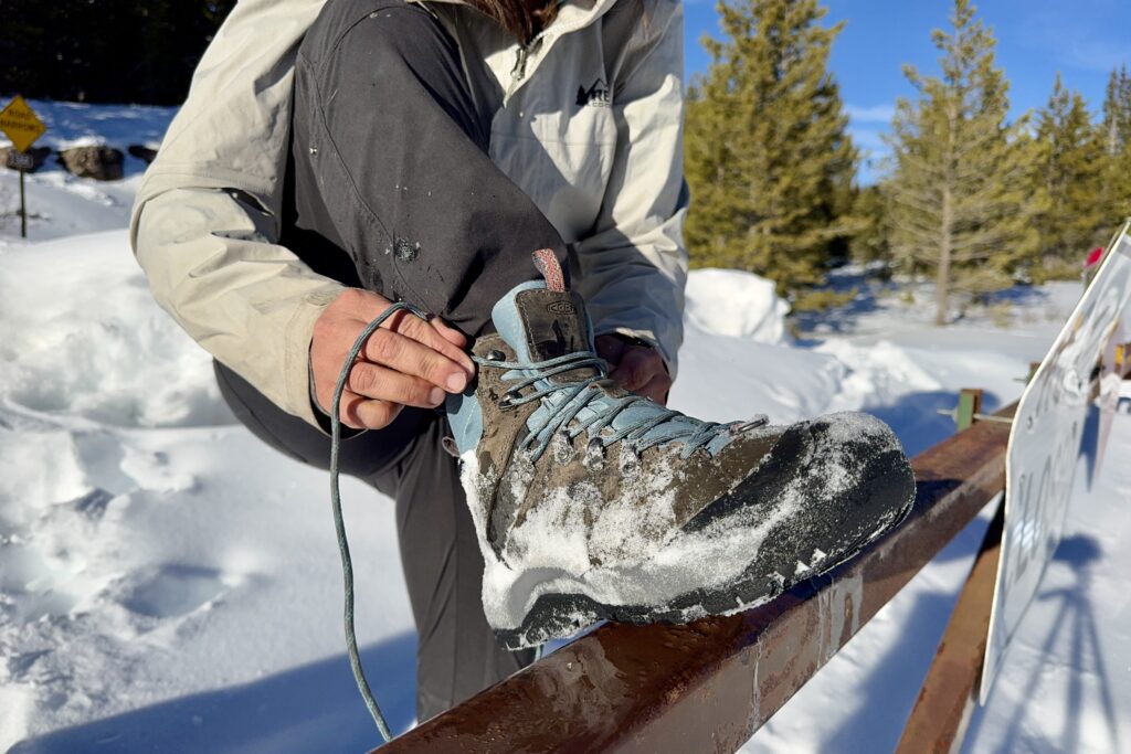 A close up of a person wearing the Keen boot and tying the laces on a gate in the snow.