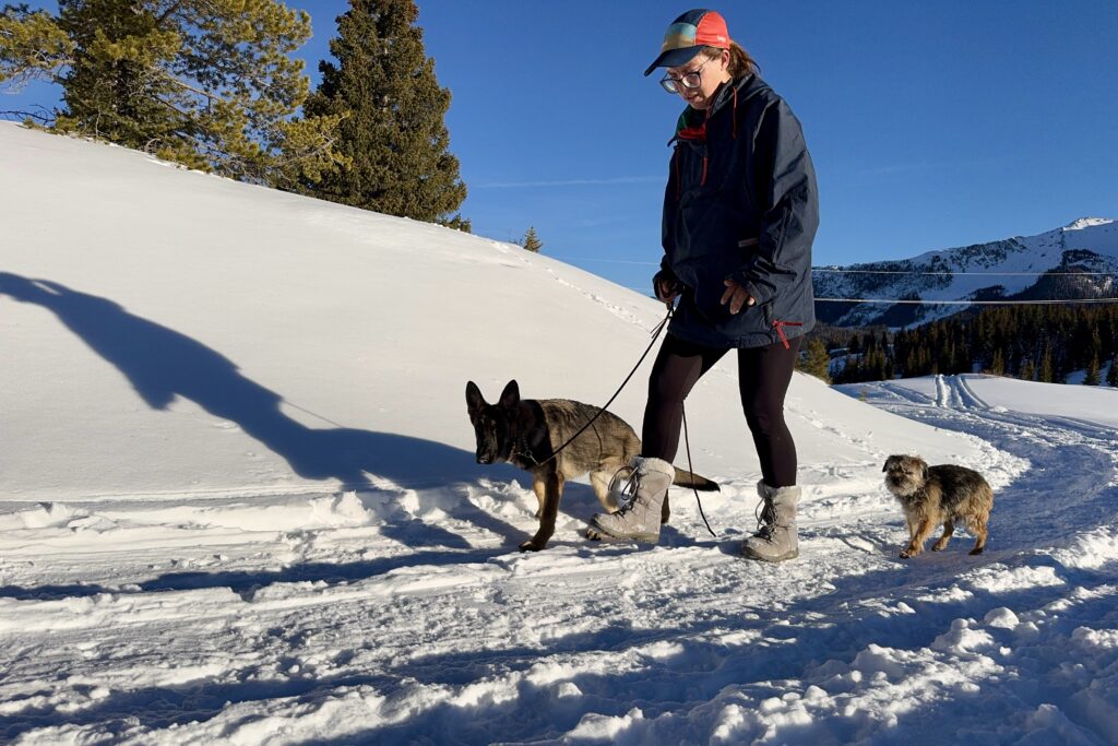 A close up of a person walking two dogs in the snow while wearing the Ice Maiden boots. There are mountains in the background.