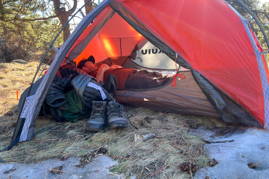 A camper relaxing inside the KUIU Storm Star 2 tent, surrounded by gear and a pair of boots outside the open vestibule, with sunlight filtering through the tent walls.