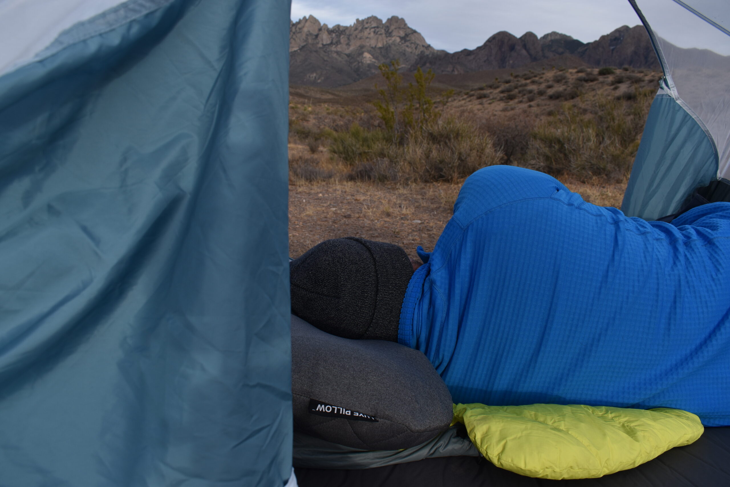 Our tester resting on his side on top of the Klymit LUXE Pillow inside an open tent facing dimly lit mountains around sunrise.