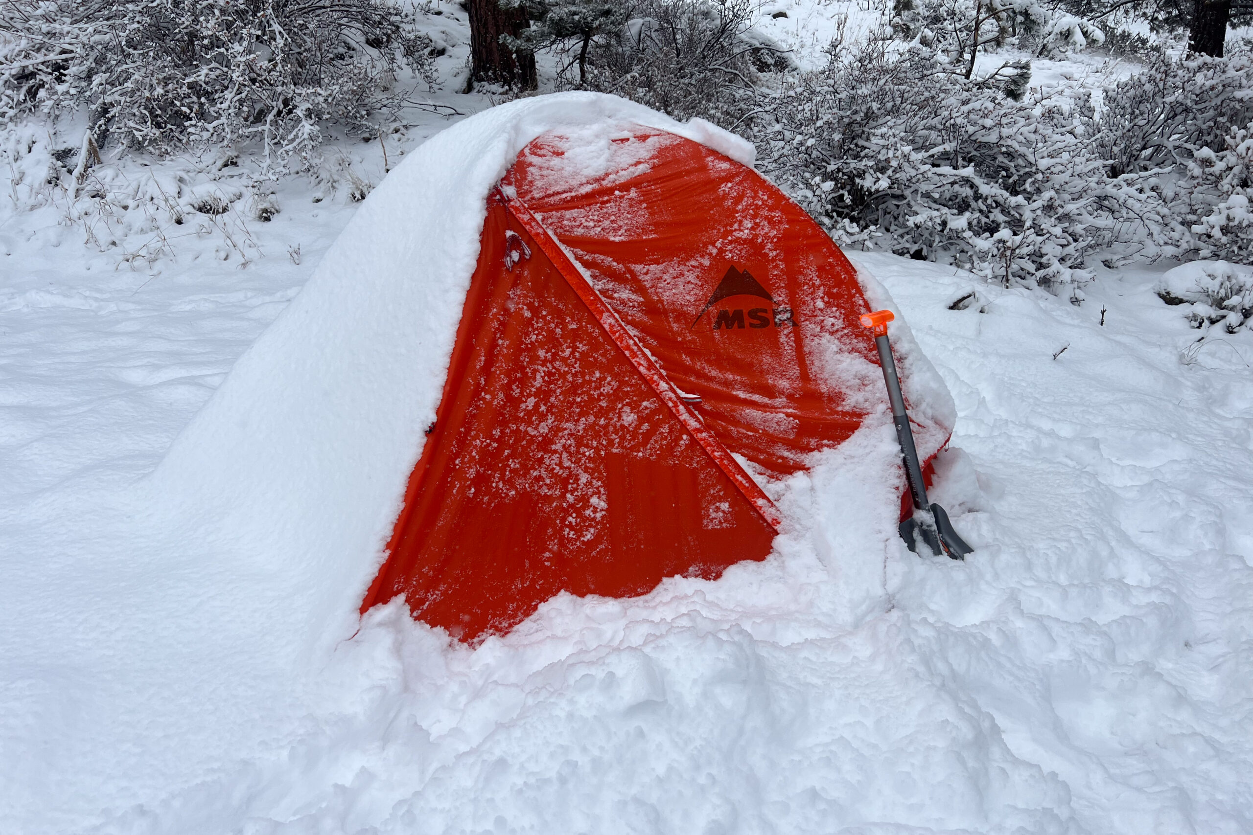 MSR Access 2 tent covered in snow with a snow shovel leaning against its entrance. The bright orange fly contrasts against the thick white snow in a forested area.