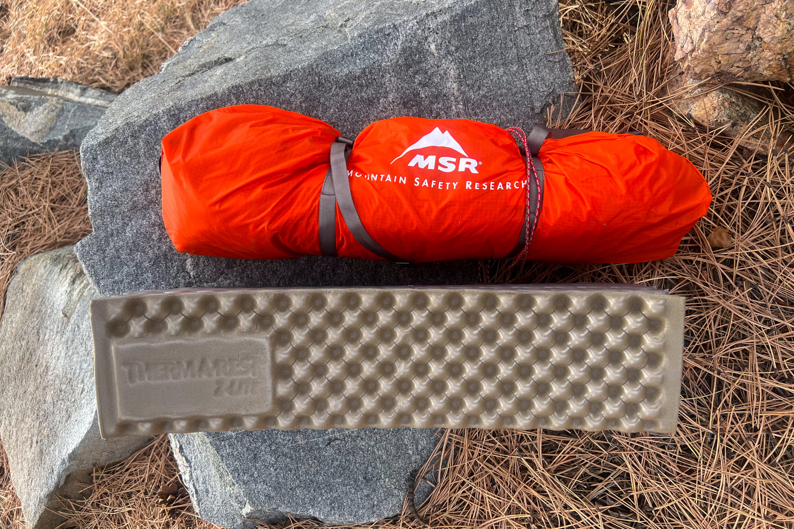 Packed MSR Access 2 tent next to a folded Therm-a-Rest ZLite sleeping pad on a rock surface with pine needles nearby.