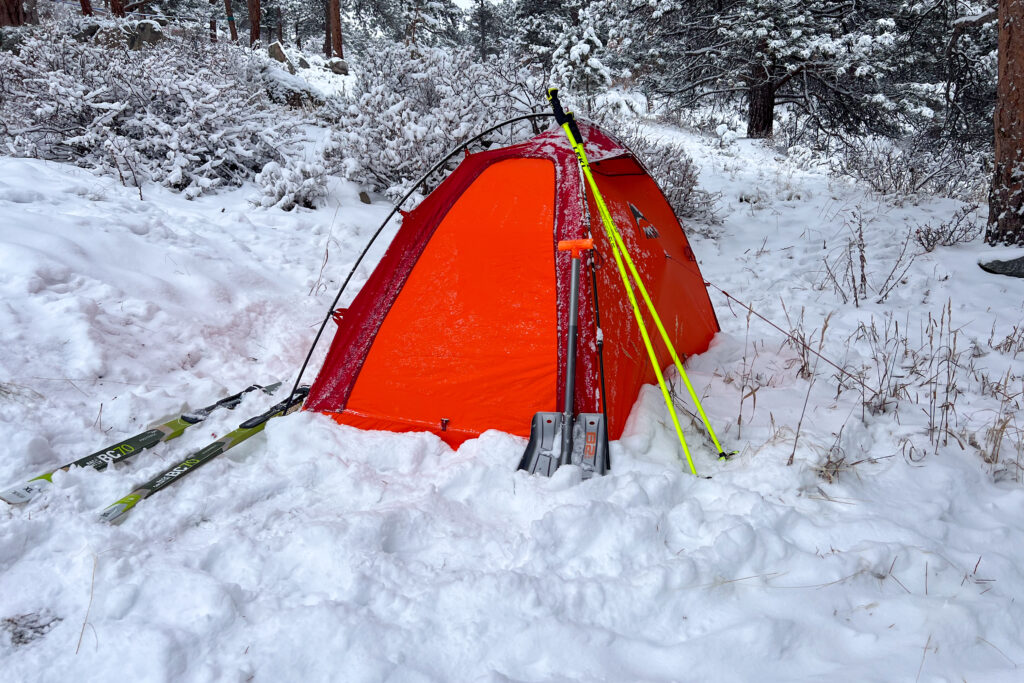 The MSR Advance Pro 2 tent pitched in the snow with skis and an avalanche shovel leaning against it, surrounded by snow-covered trees.