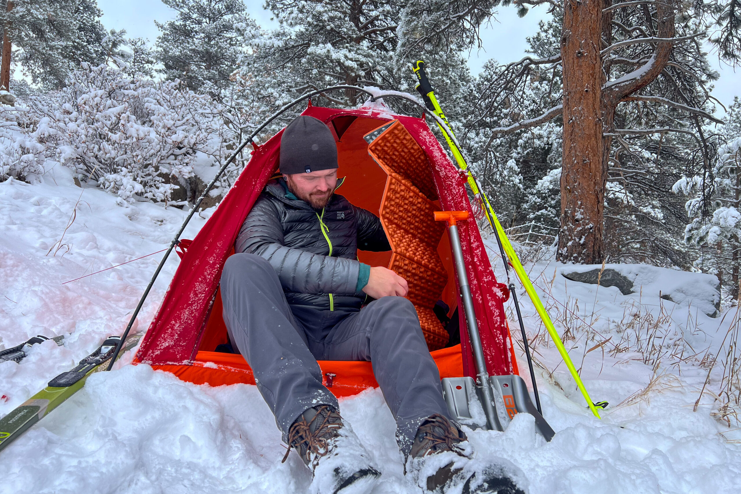 A person sitting at the entrance of an MSR Advance Pro 2 tent, wearing a puffy jacket and preparing gear inside the tent, with snow-covered skis and trekking poles visible outside.