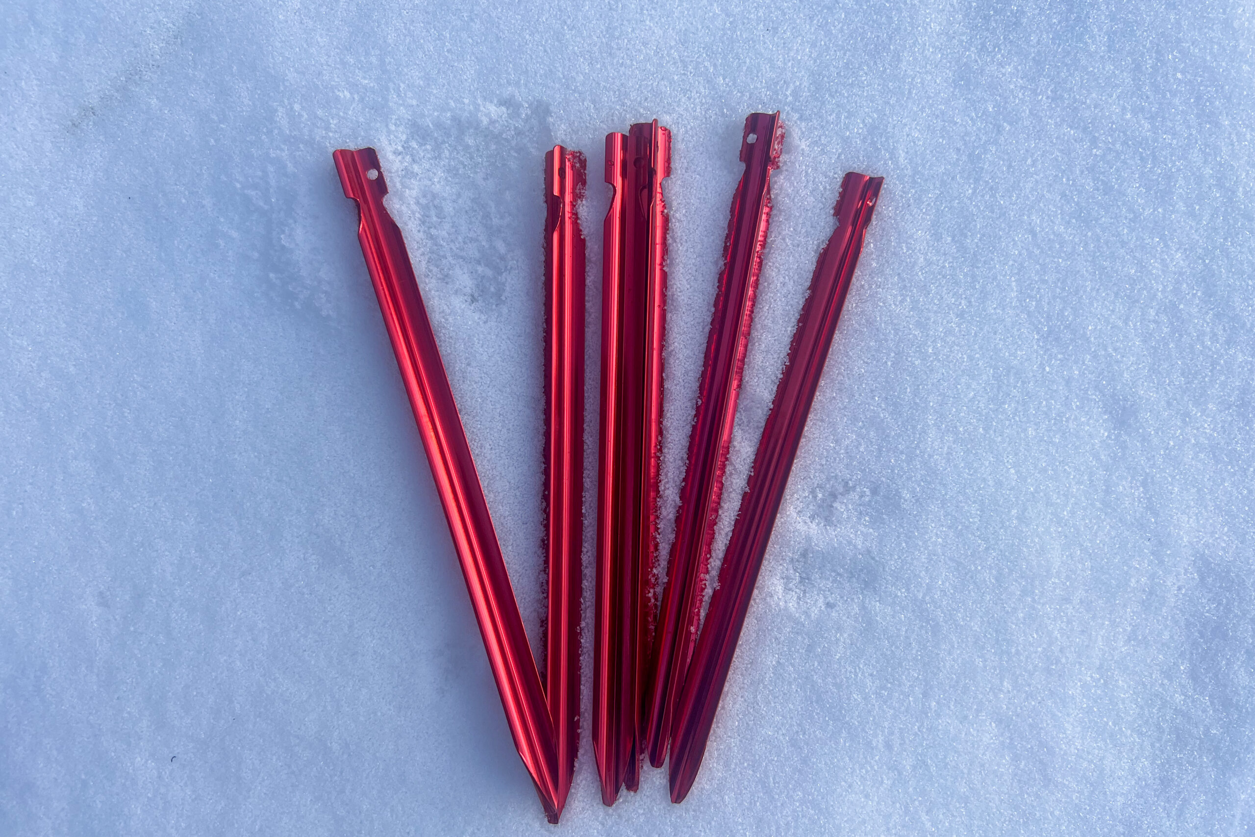 A set of five red MSR tent stakes lying on snow, showcasing their pointed design and lightweight construction.