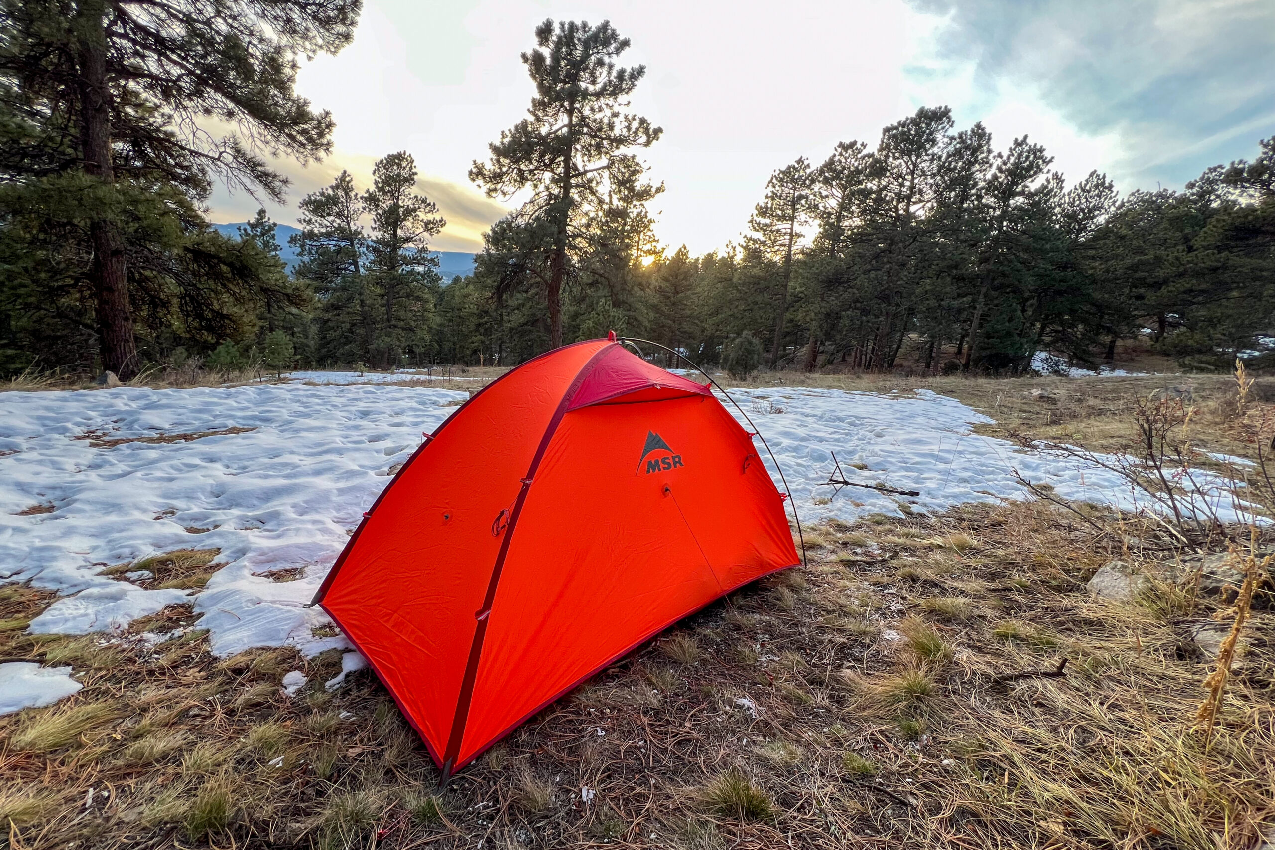 A red MSR Advance Pro 2 tent pitched on a snowy field surrounded by pine trees, with the sunset in the background.