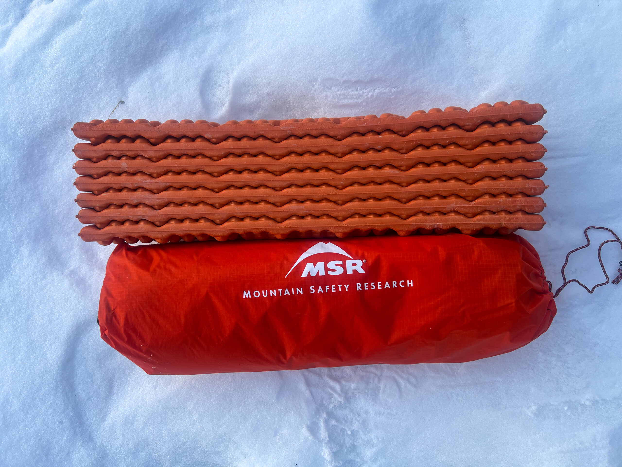 The MSR Advance Pro Tent packed into it's stuff sack and laying in the snow next to a foam sleeping mat.