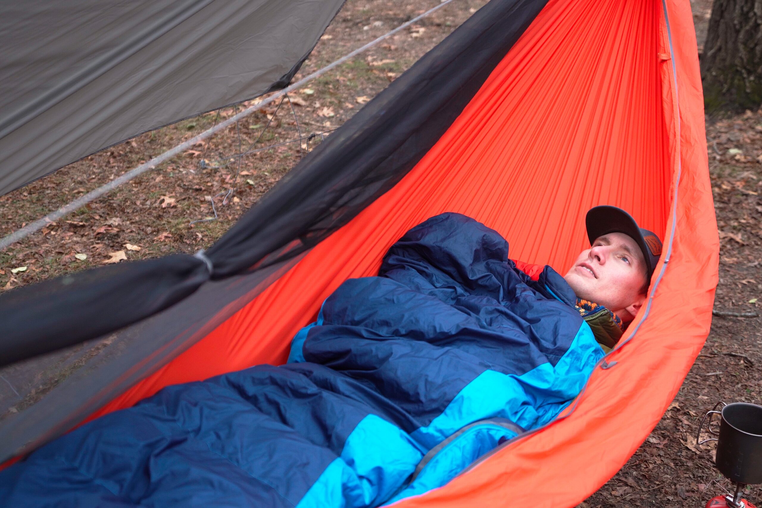 A man in a blue sleeping bag lays back in an orange hammock with the bug net tied back