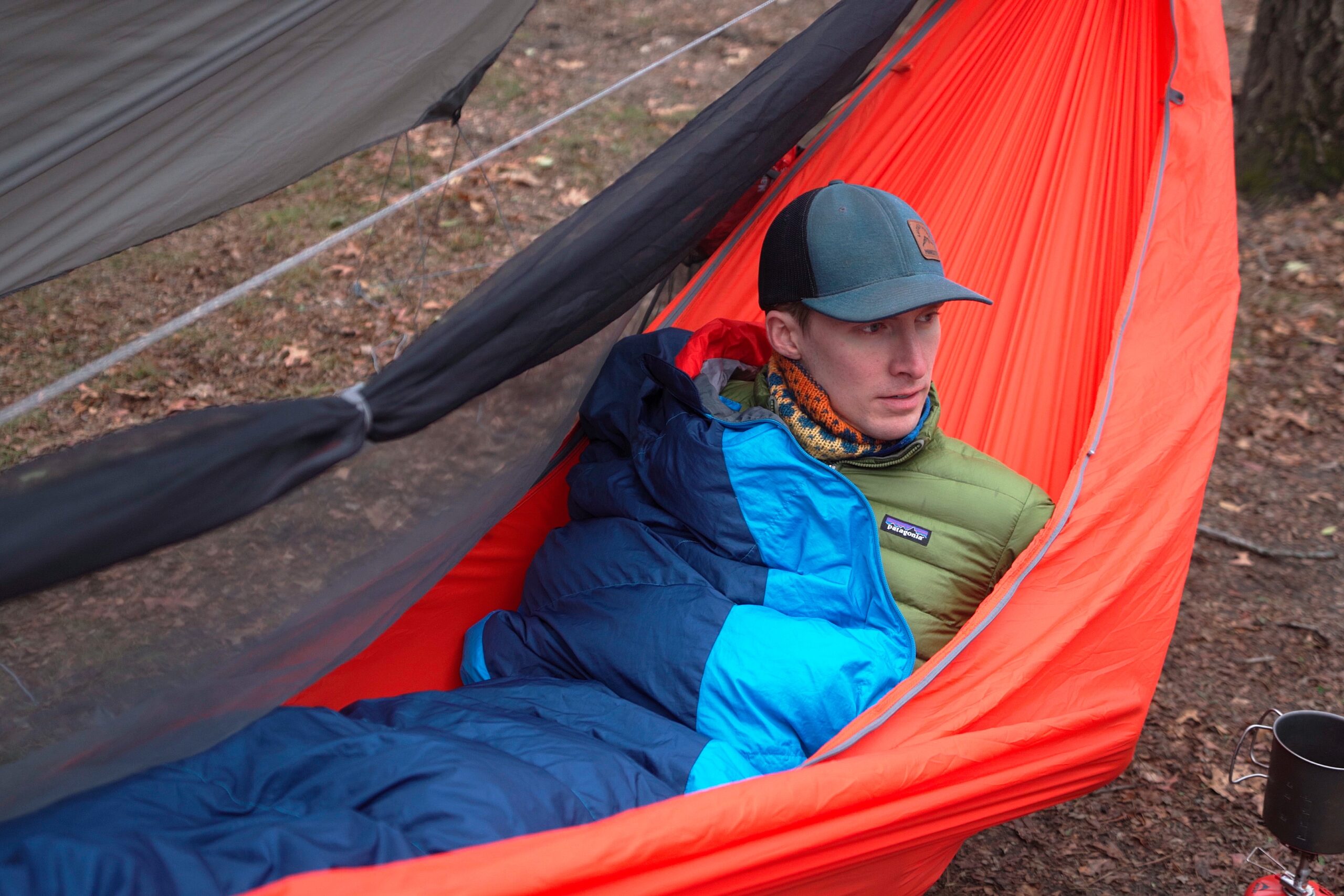 a man in a blue hat in a blue sleeping bag sits up in an orange hammock with the bug netting rolled back