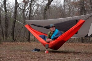 A man in a green coat sits in an orange hammock in a forest