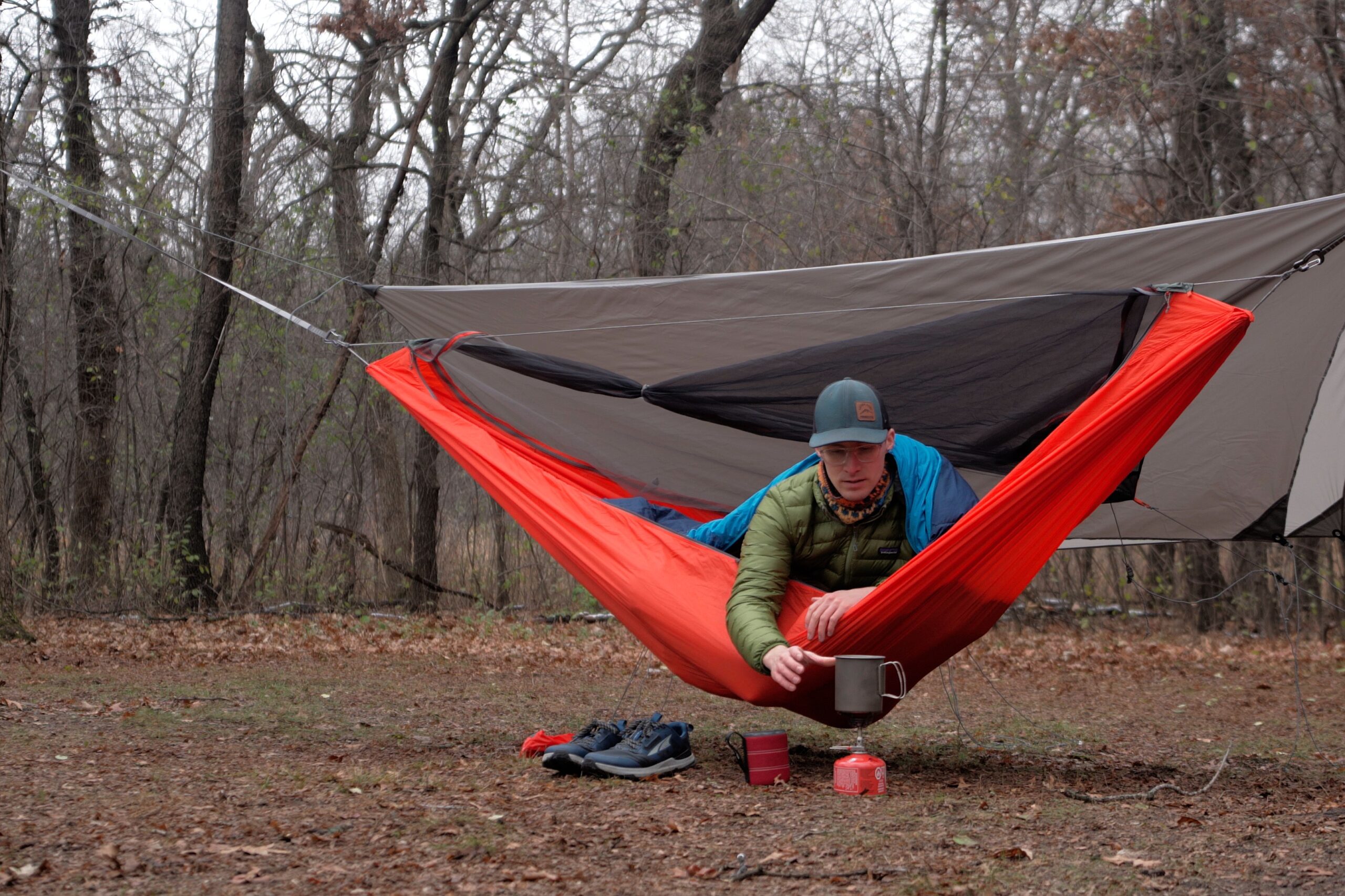 A man in a green coat sits in an orange hammock in a forest cooking on a backpacking stove