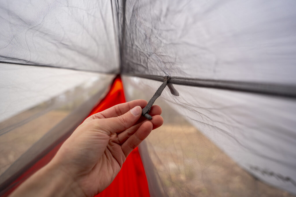 A hand holds a toggle on the interior of a hammock bug netting