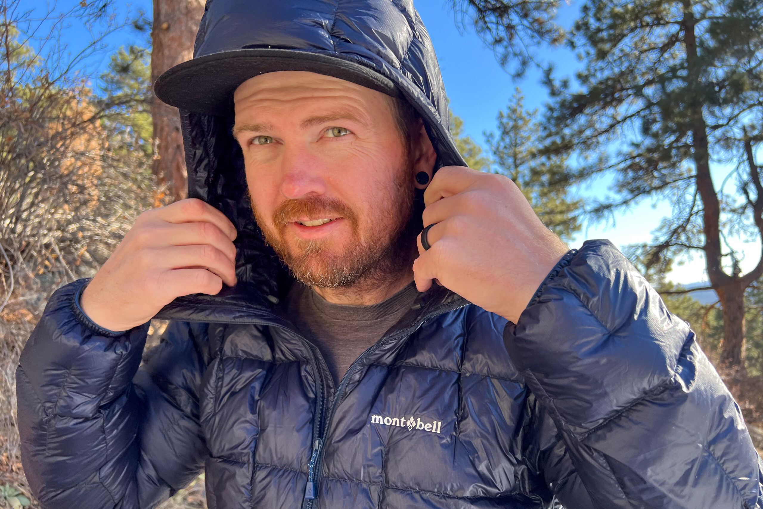 A close-up of a person wearing the MontBell Plasma 1000 Alpine Down Parka, holding the hood with both hands in a winter forest setting.