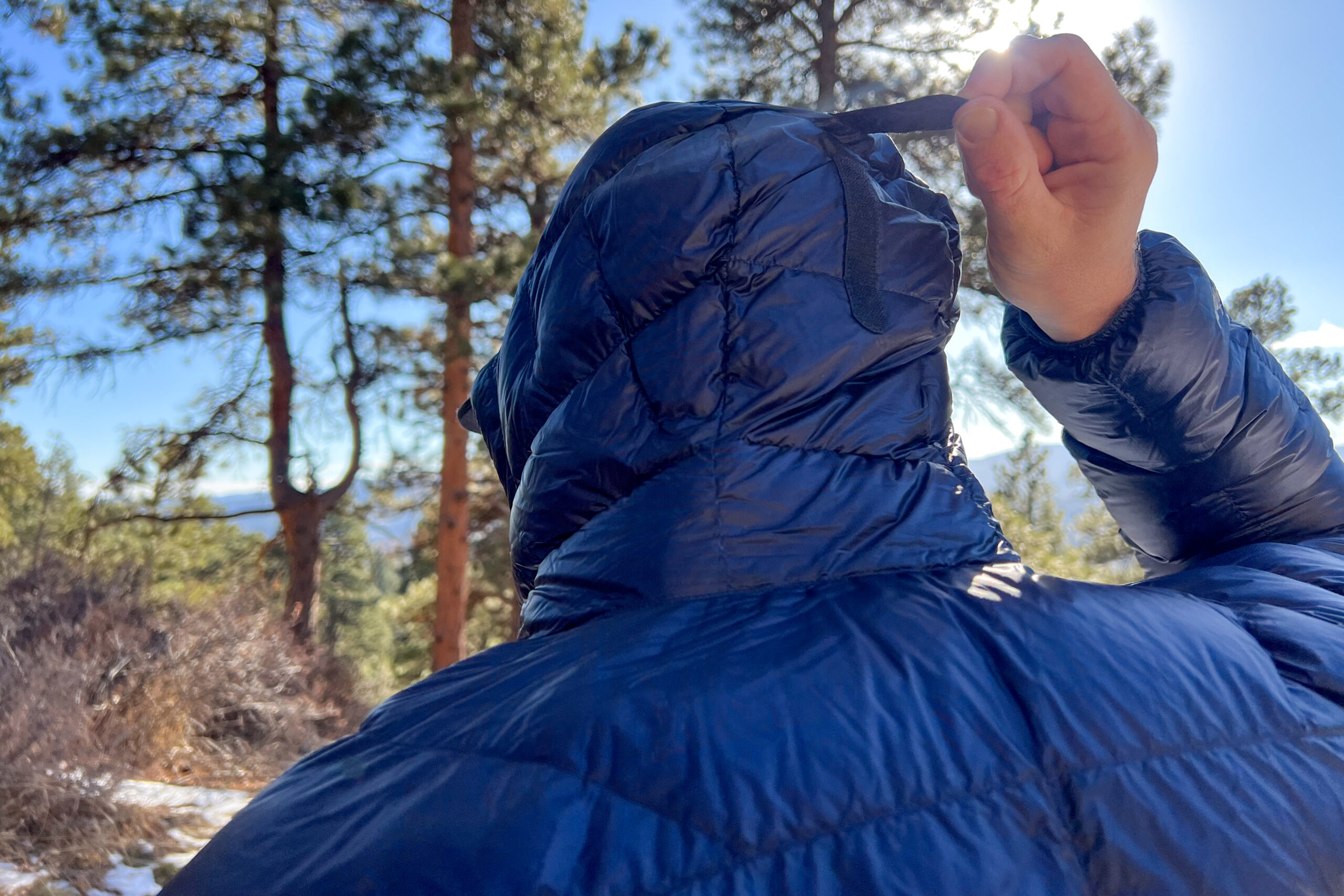 Close-up of the MontBell Plasma 1000 Alpine Down Parka's hood being adjusted with a rear pull cord against a sunny forest backdrop.