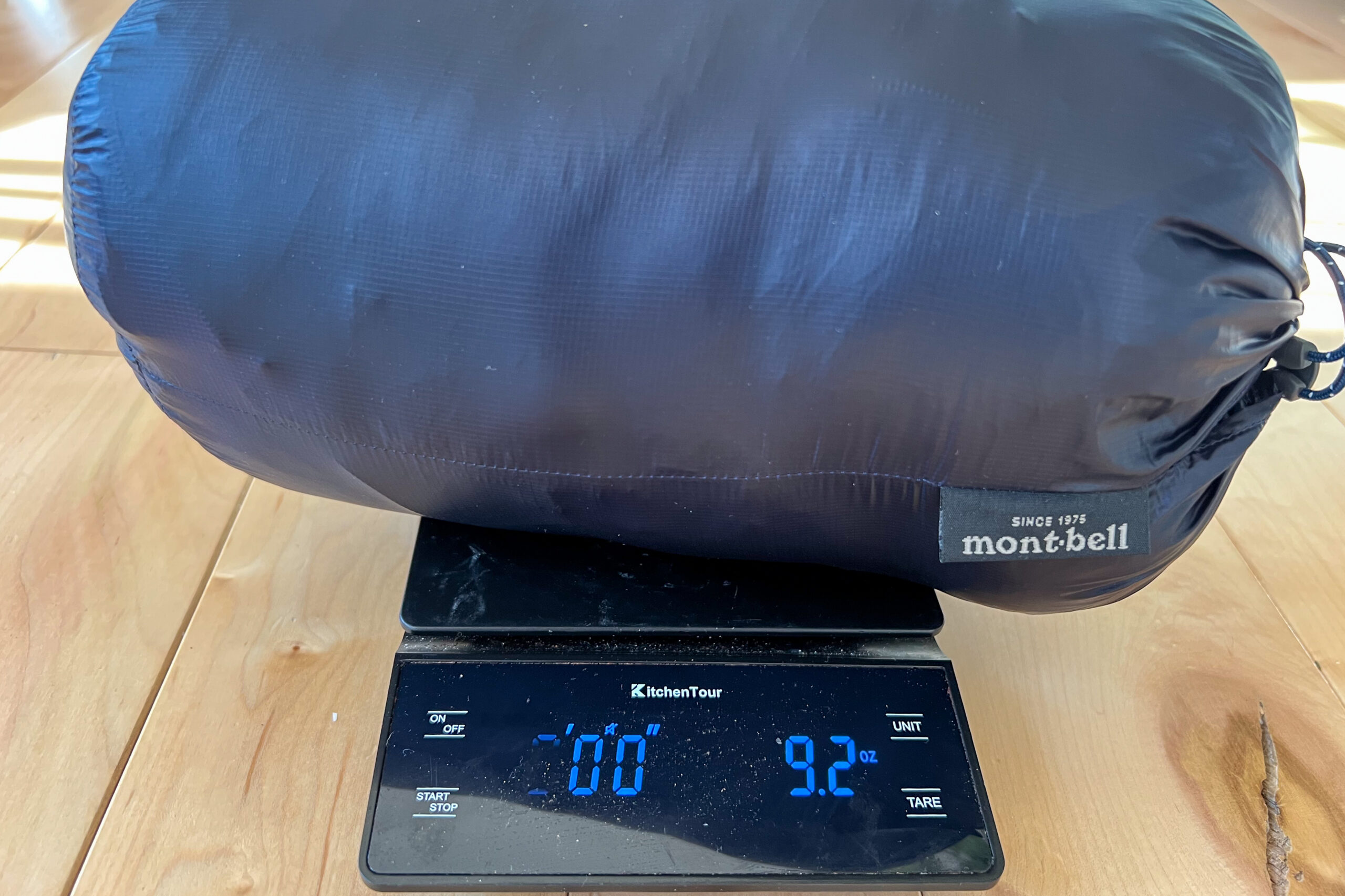 The MontBell Plasma 1000 Alpine Down Parka packed in its stuff sack on a digital scale, displaying a weight of 9.2 ounces.