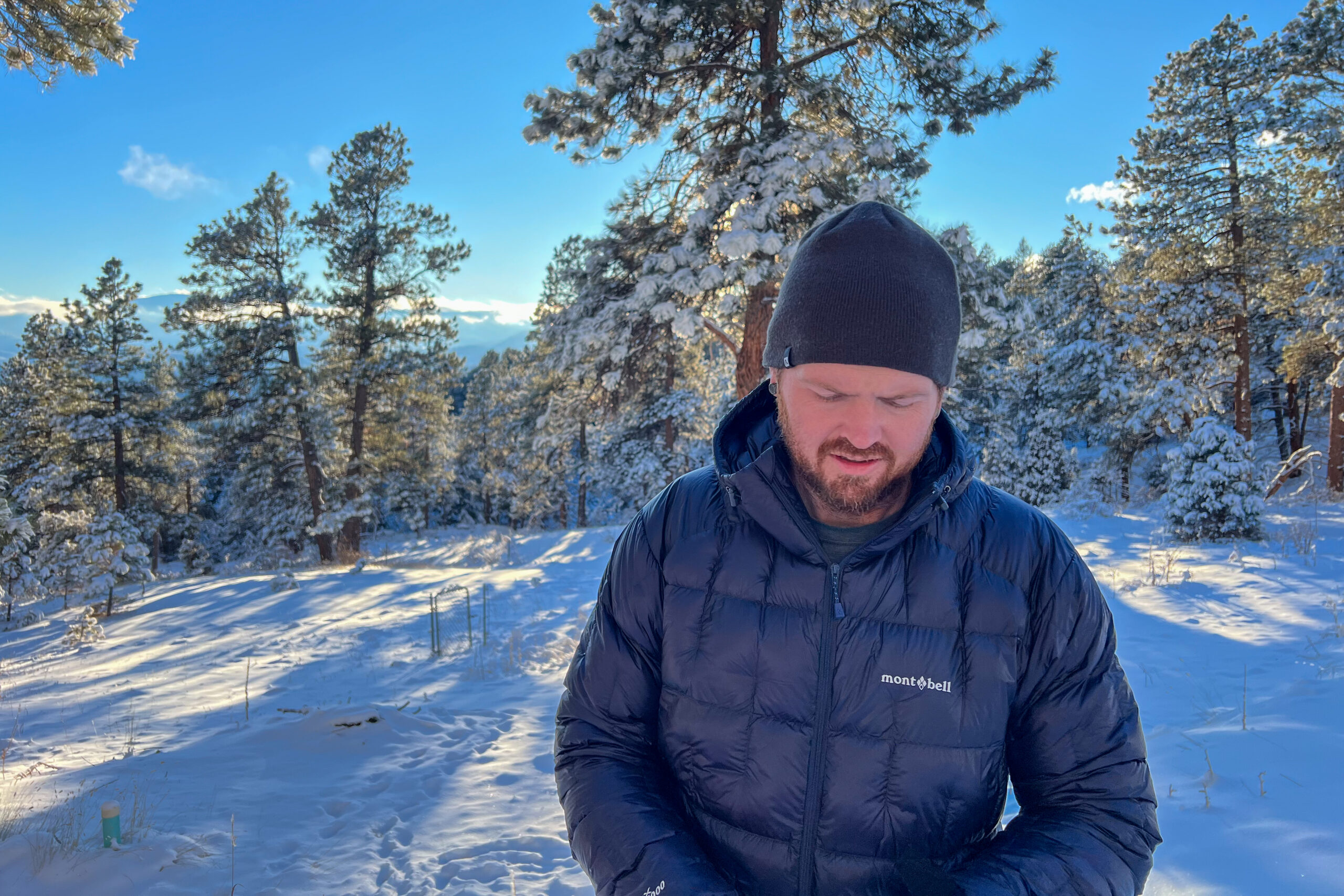 A person wearing the MontBell Plasma 1000 Alpine Down Parka, standing in a snowy forest and looking down at their jacket, surrounded by snow-covered trees.