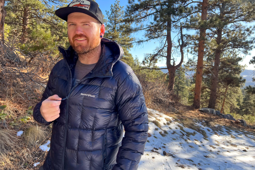 A person wearing the MontBell Plasma 1000 Alpine Down Parka, standing outdoors on a snowy trail while adjusting the front zipper.