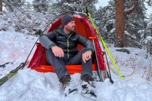 A person sits in a red tent pitched on snow, wearing a lightweight down jacket with snow-covered boots.