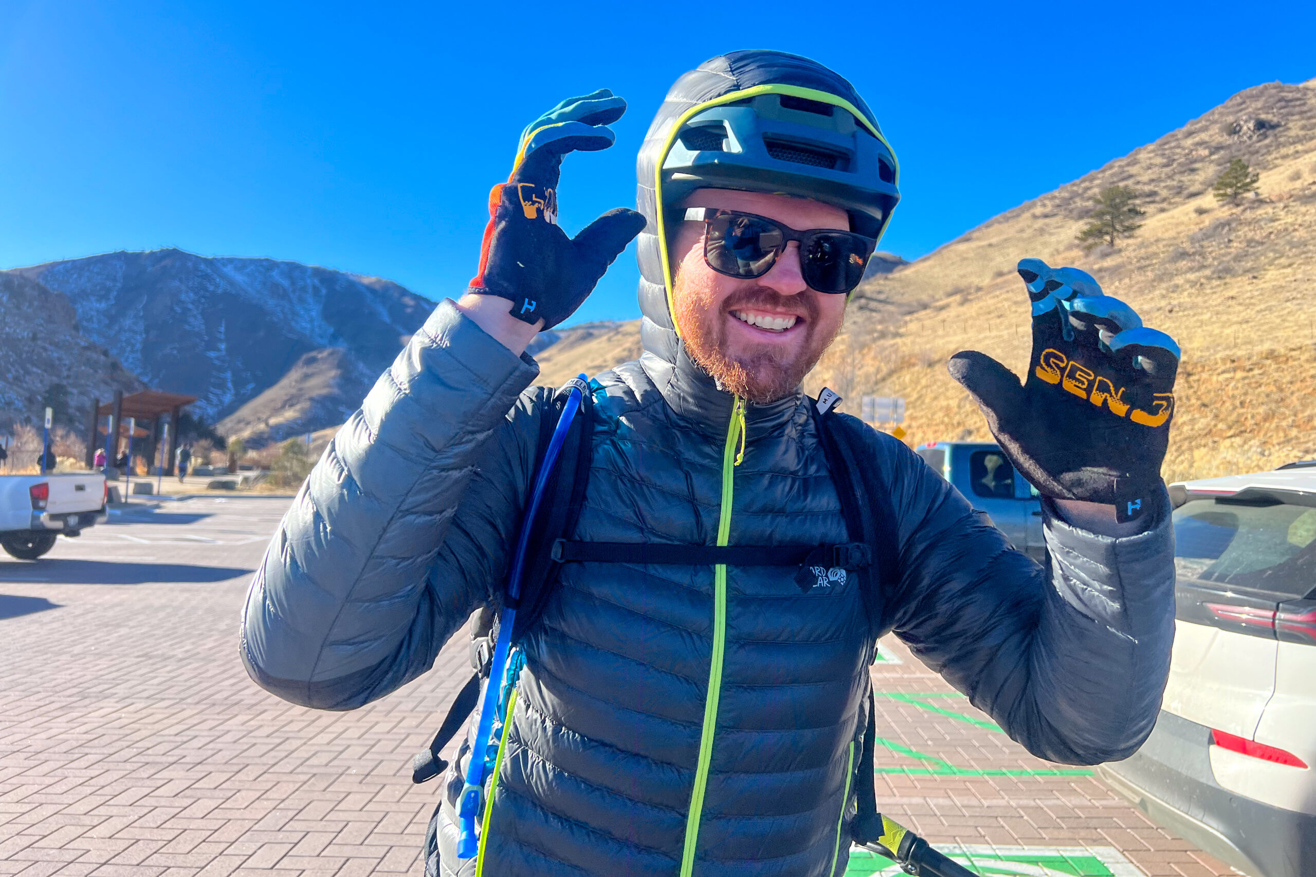 A person wearing a helmet, gloves, and a down jacket stands smiling in a parking lot with mountain scenery.