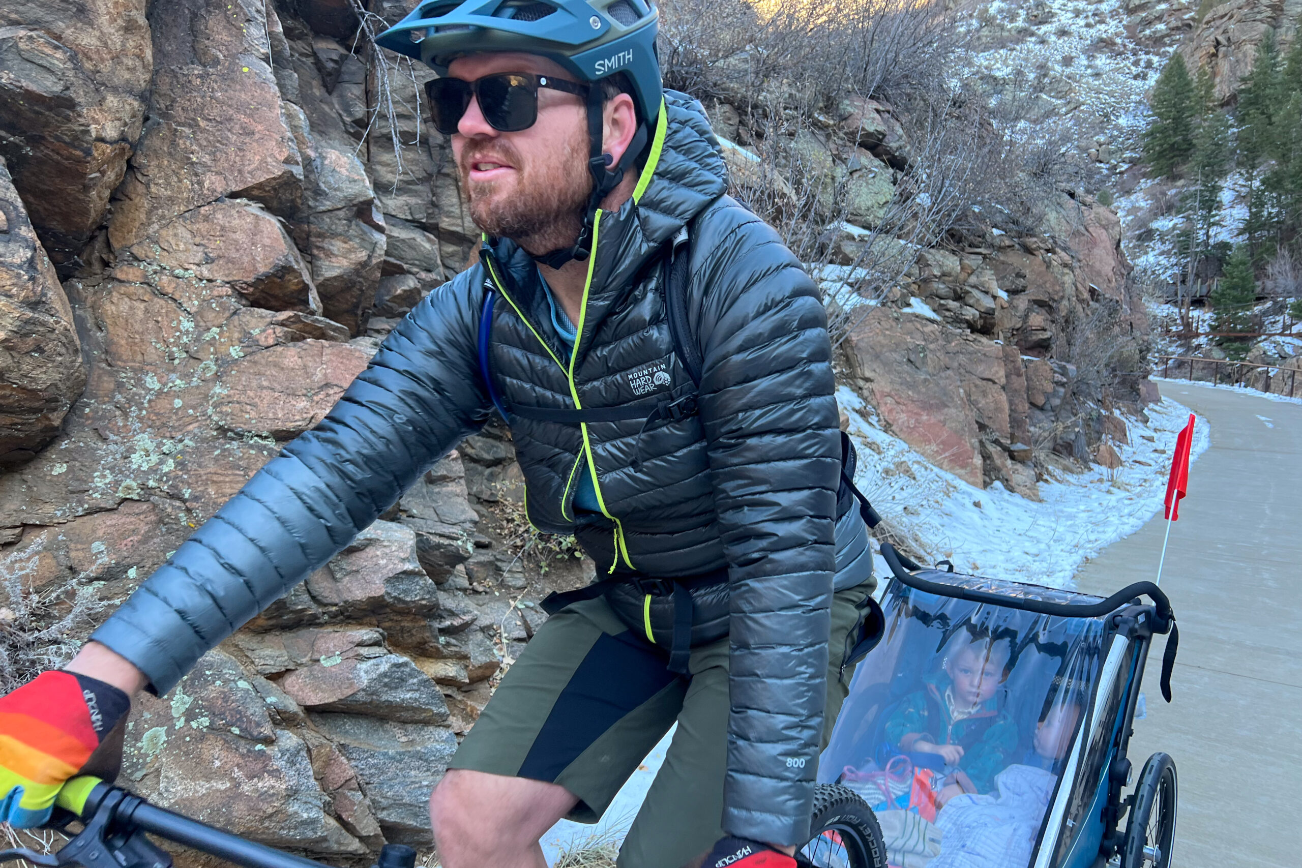 A person in a helmet and Ghost Whisperer/2 Hoody rides a bike while pulling a child trailer, with rocky cliffs in the background.