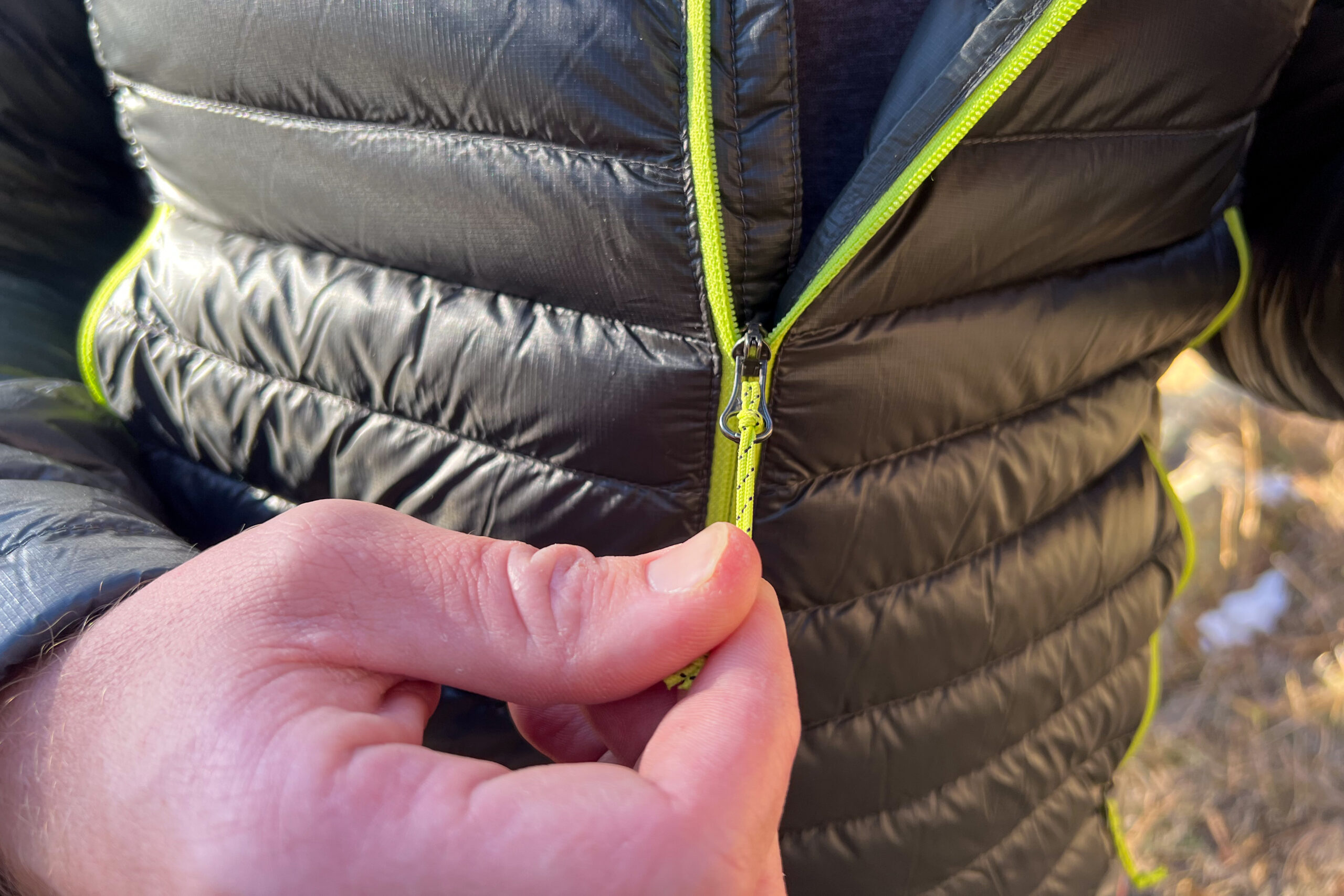 A close-up of a person's hand holding the front zipper pull on a lightweight down jacket.